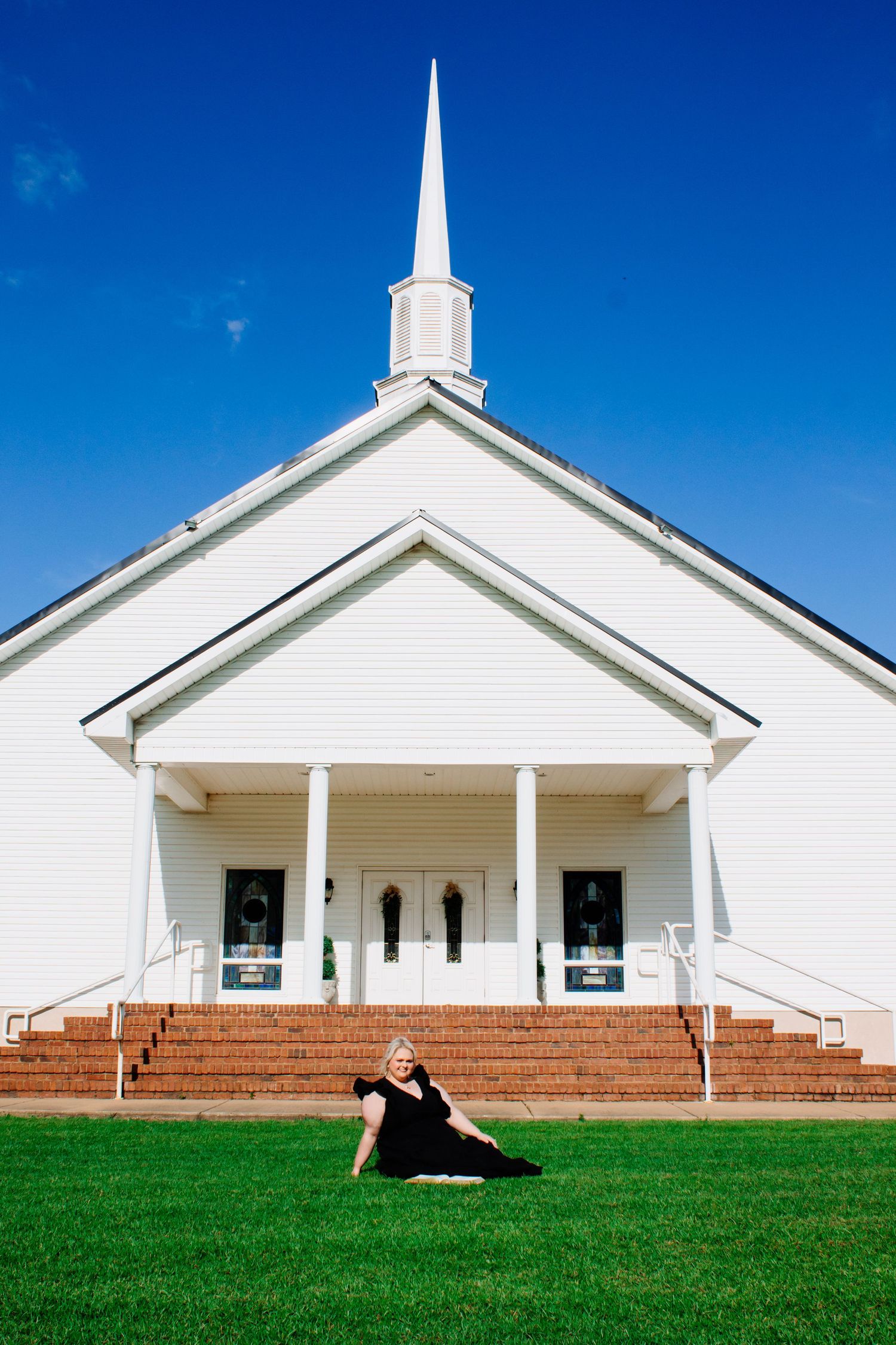 Blonde woman in black dress sitting on green lawn in front of white church with tall steeple.