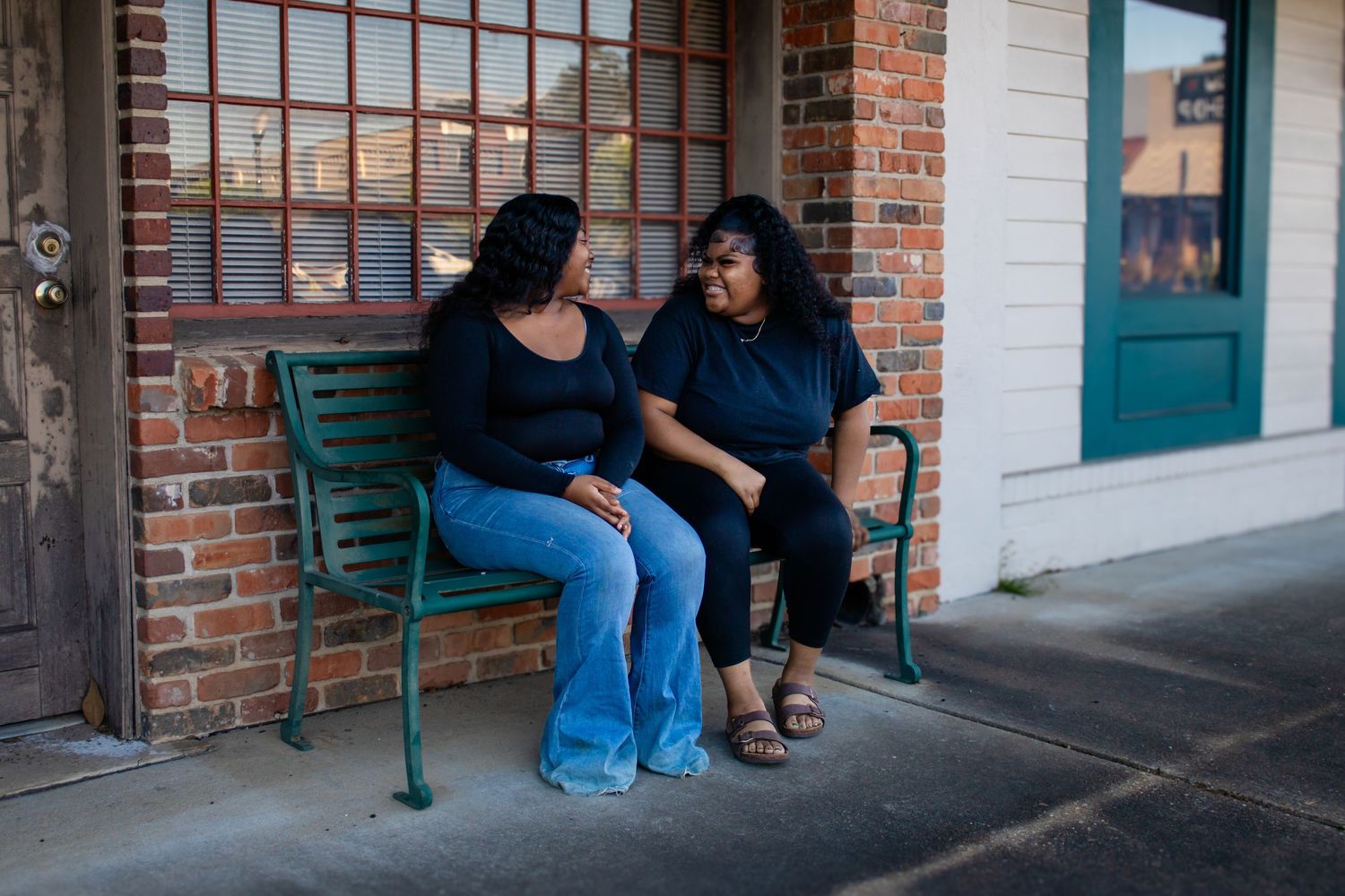 Two young Black women in black outfits sitting together on a teal bench outside a brick building.