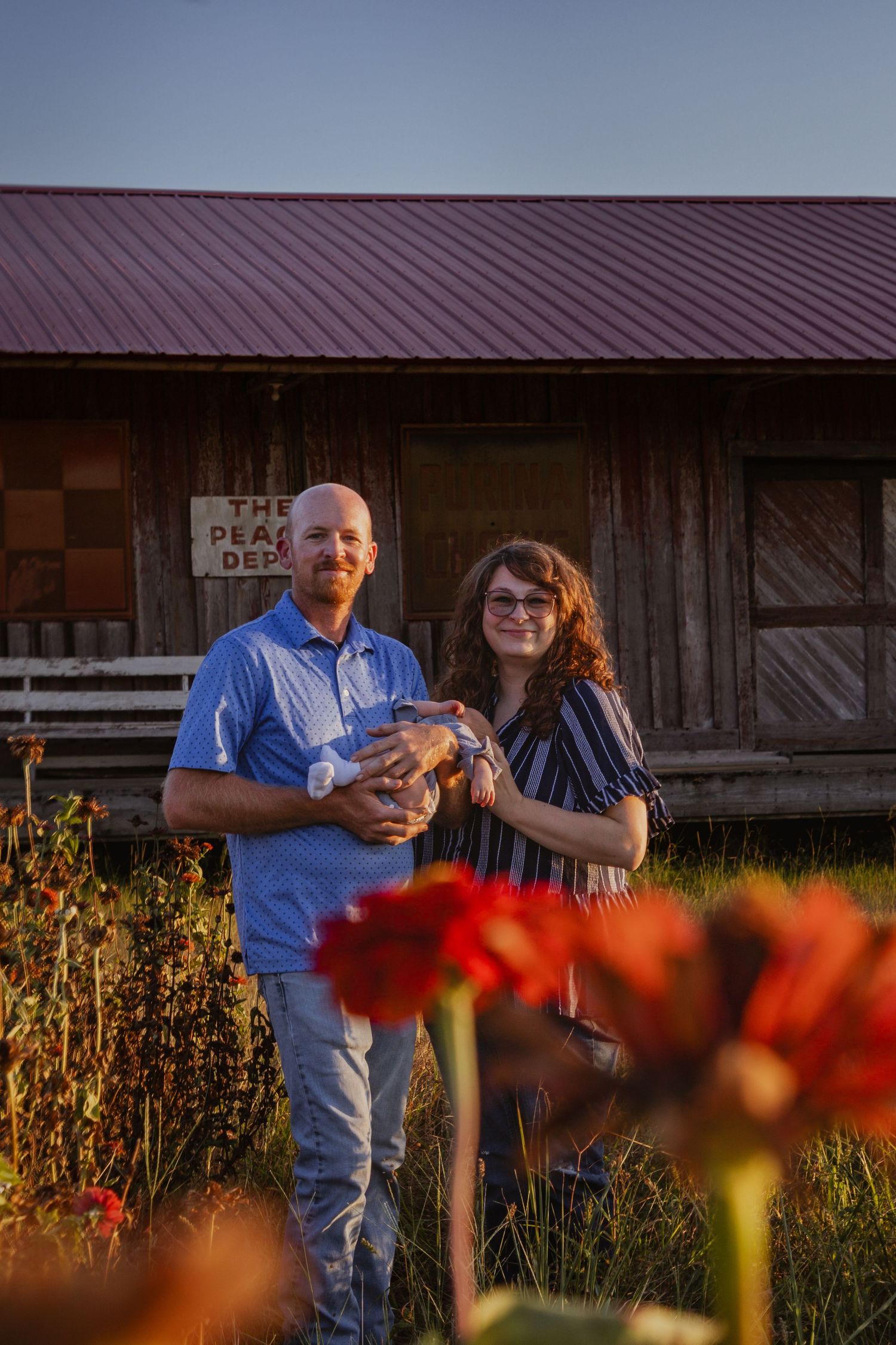 Smiling couple holding newborn near rustic barn with red roof and red flowers in foreground.