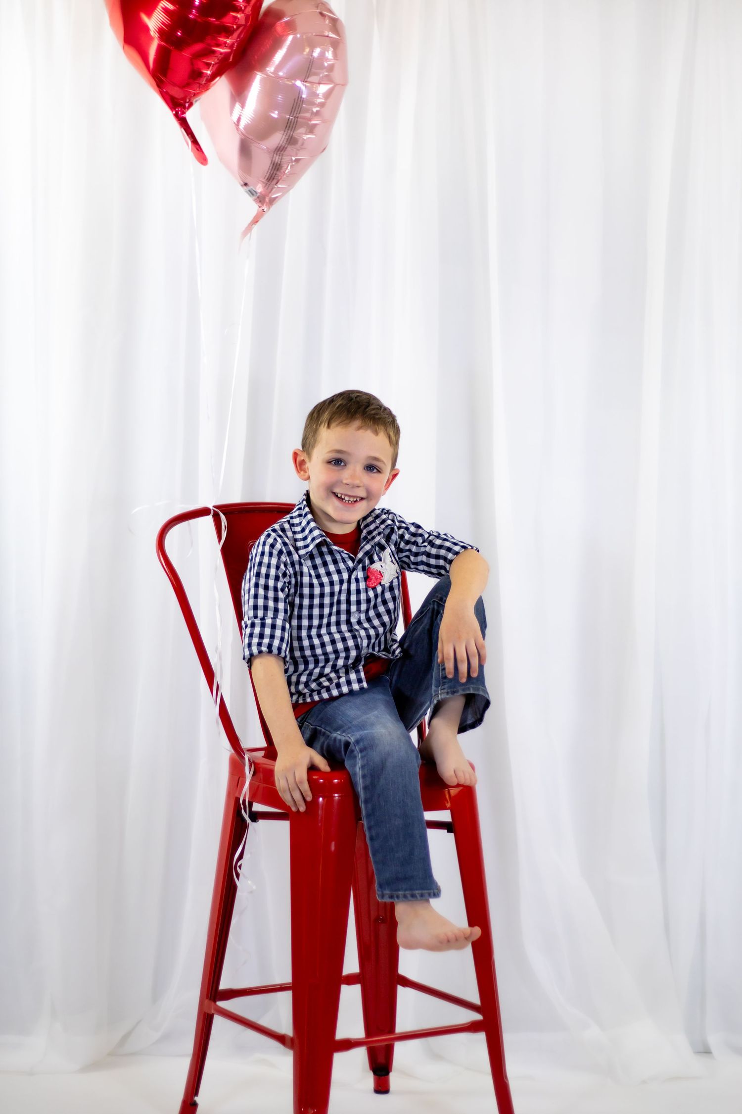 Smiling young boy in plaid shirt sits on red metal chair with heart-shaped balloons behind him.