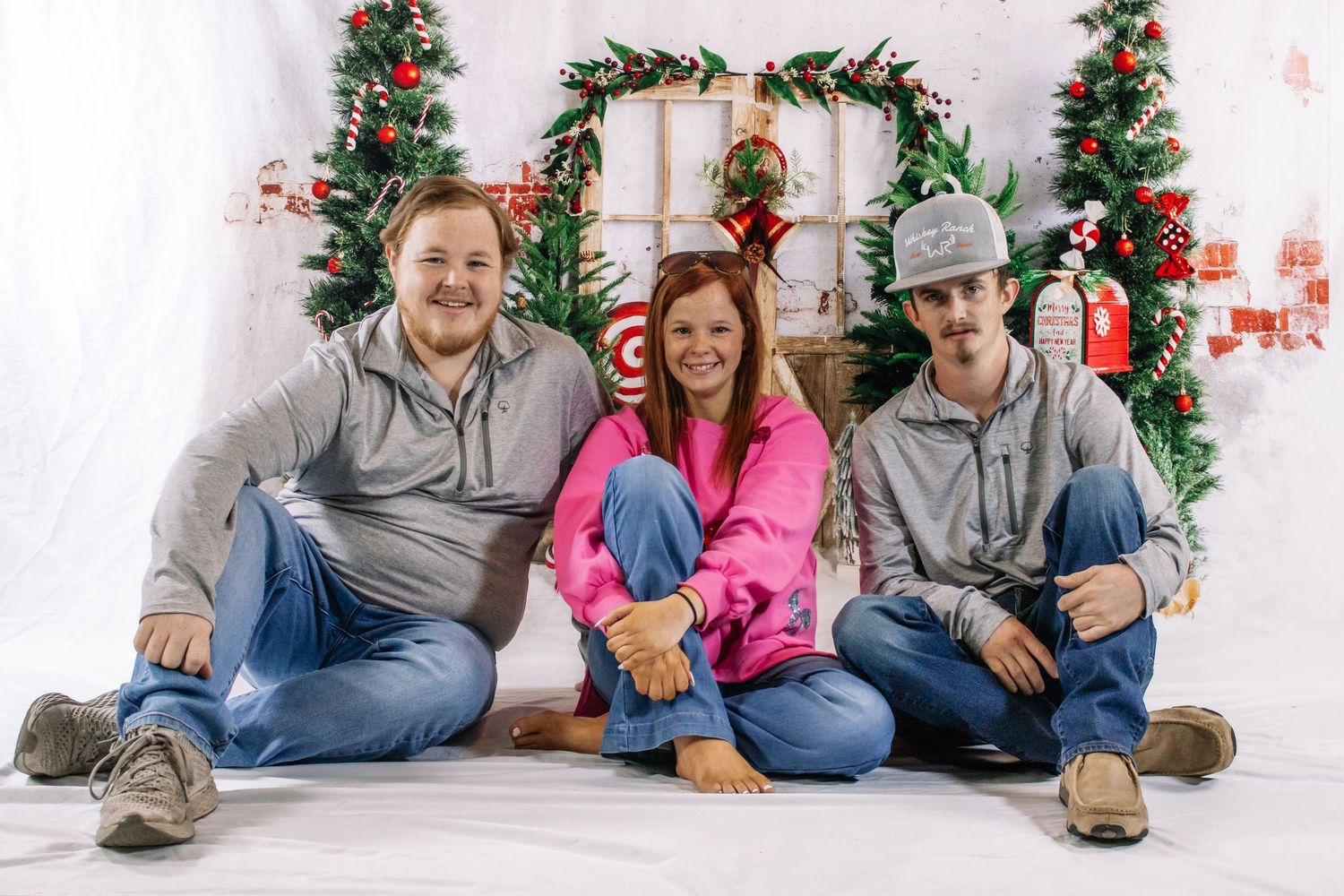 Three siblings in casual attire sitting together in front of Christmas holiday photo backdrop.