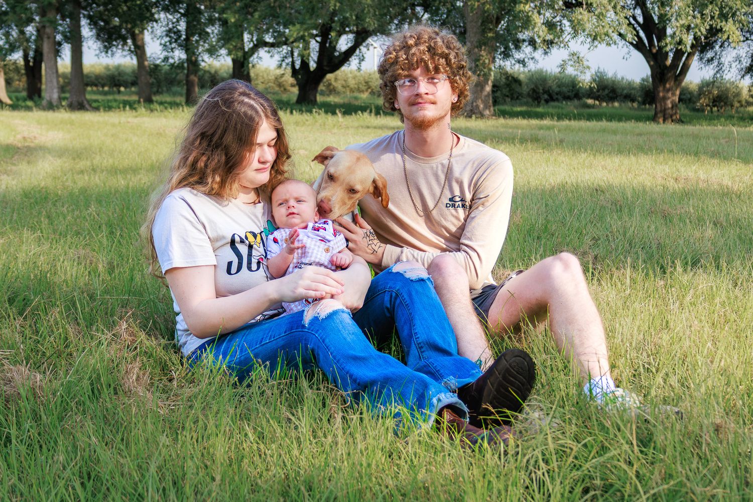 Young couple sitting in grass with newborn baby and dog on sunny day outdoors.