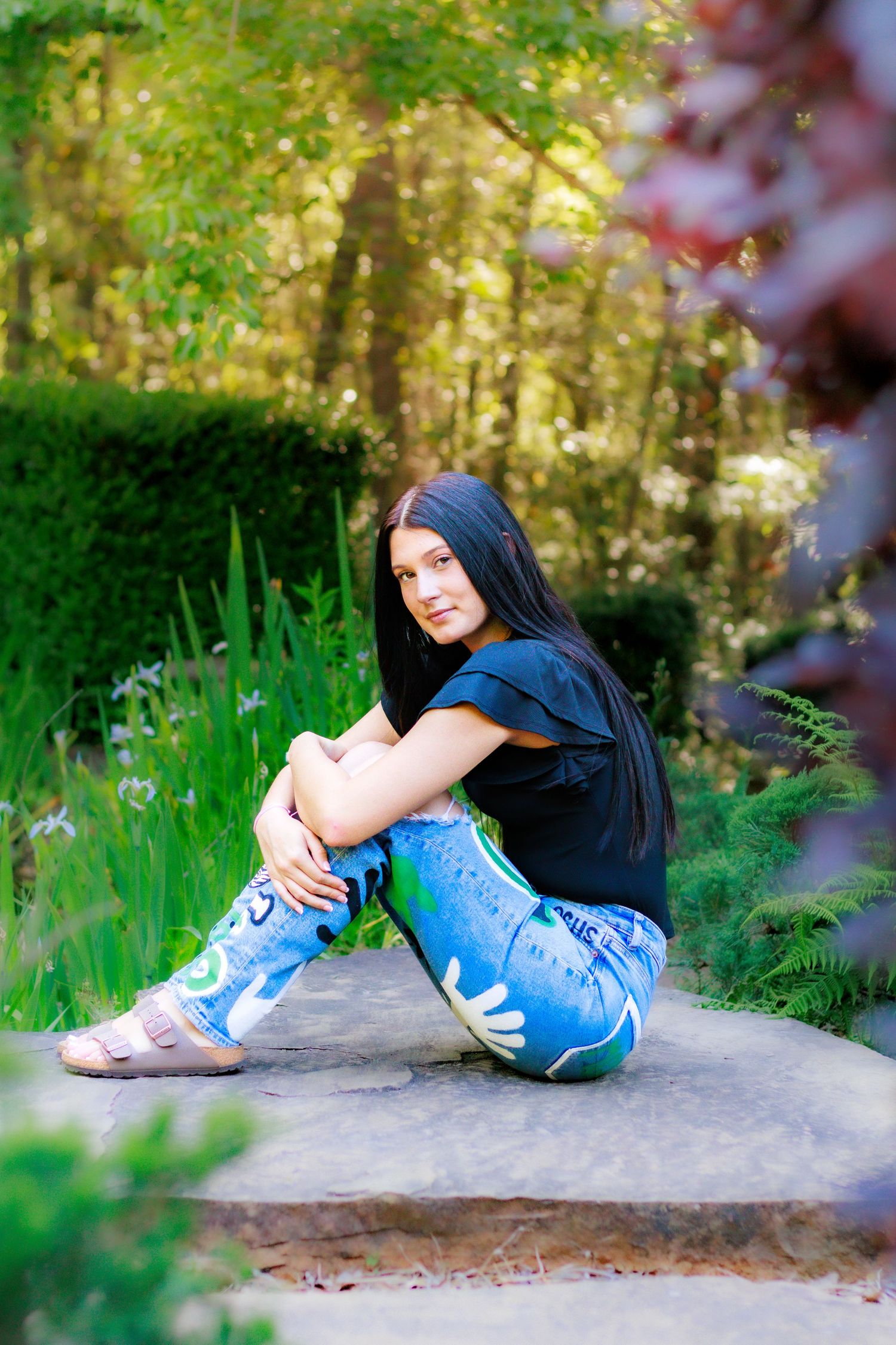 Dark-haired young woman sits on stone step in lush green garden, wearing black top and artistic painted blue jeans.