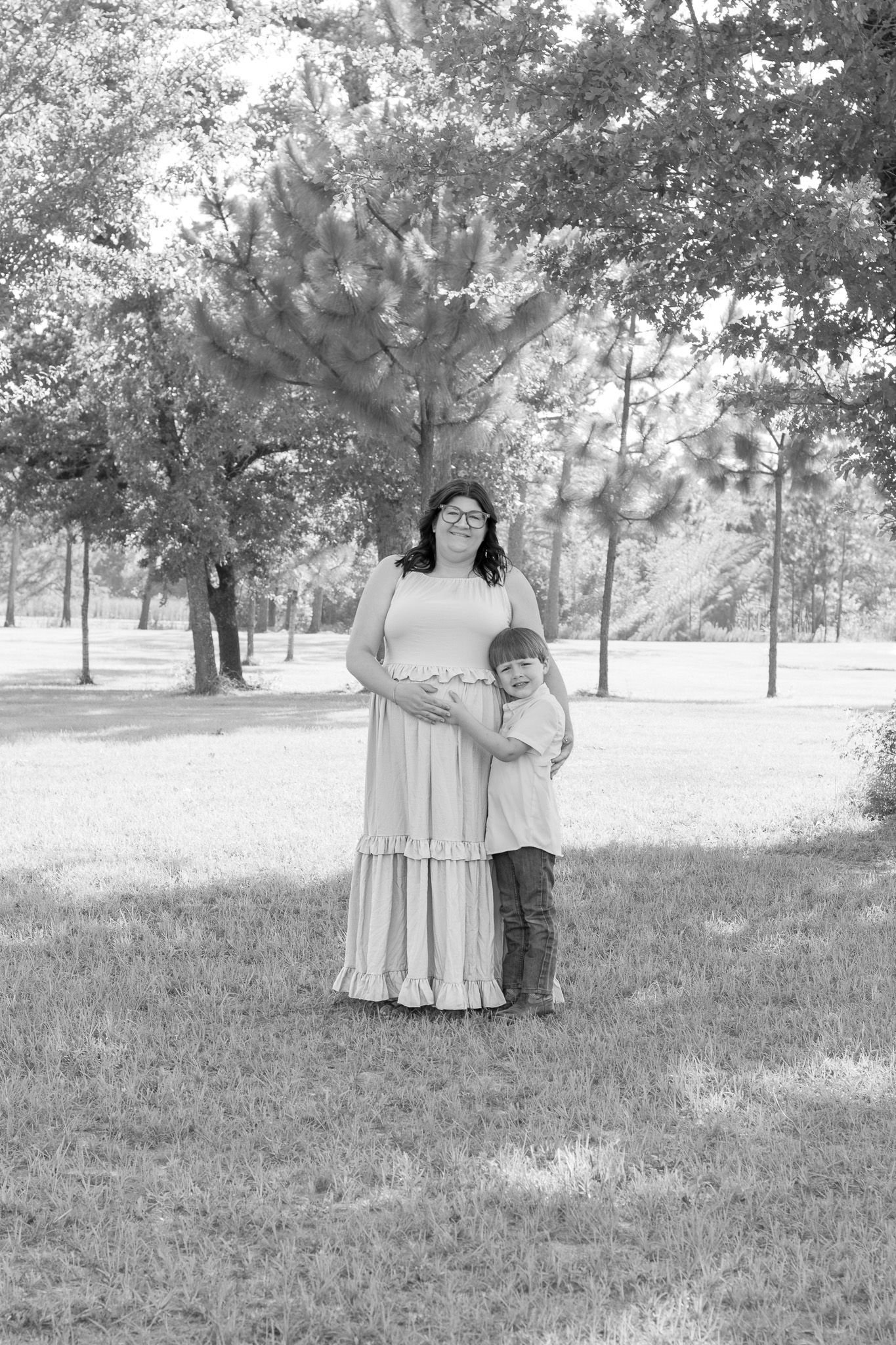 Pregnant woman in a long dress holds a child in a lush green park, captured in black and white.