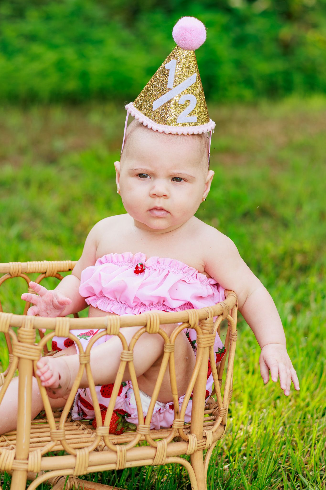 Baby in gold glitter half birthday party hat sits in wicker chair on green grass outdoors.