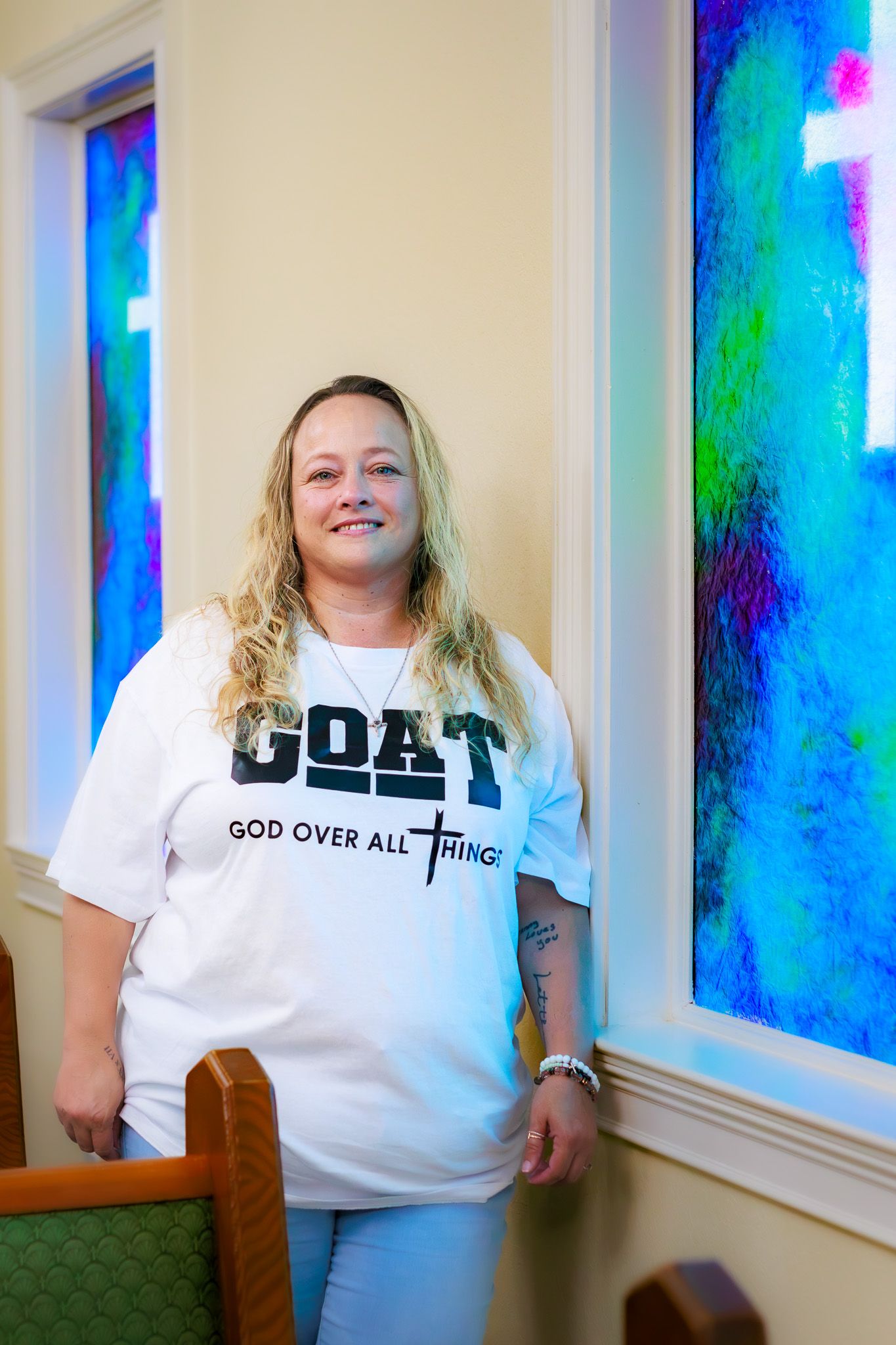 Woman in GOAT God Over All Things white tee smiling inside church with colorful stained glass windows.