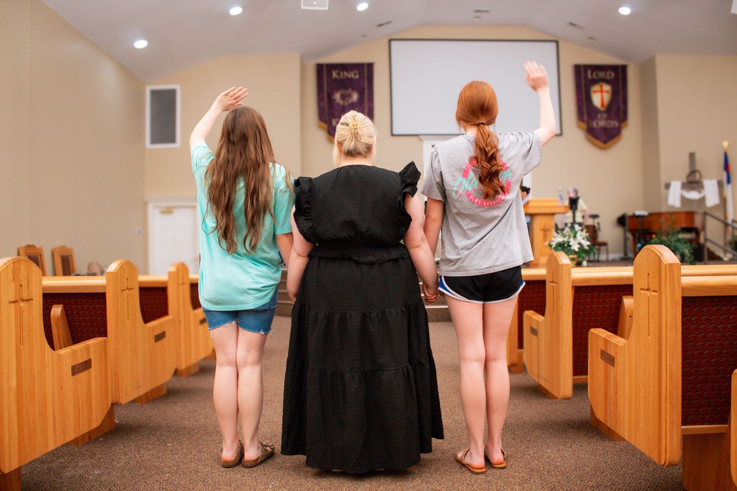 Three women standing at the altar of a church with arms raised in worship, viewed from behind.