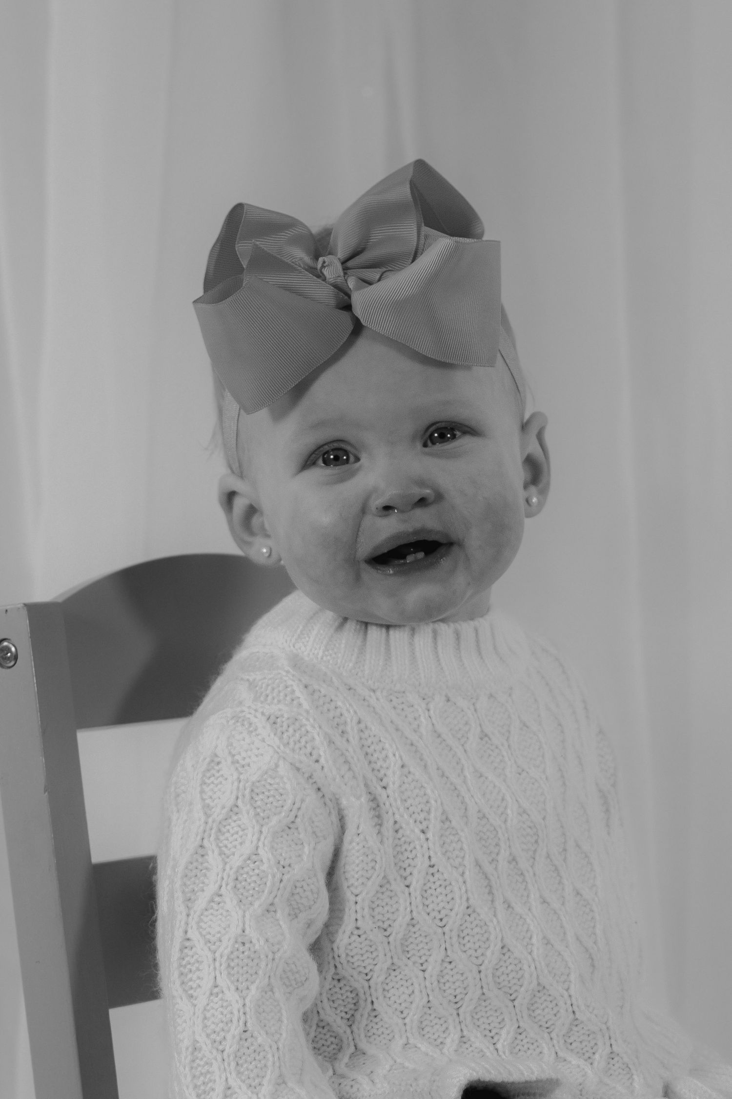 Black and white photo of a smiling baby girl in cable knit sweater wearing a large bow headband.