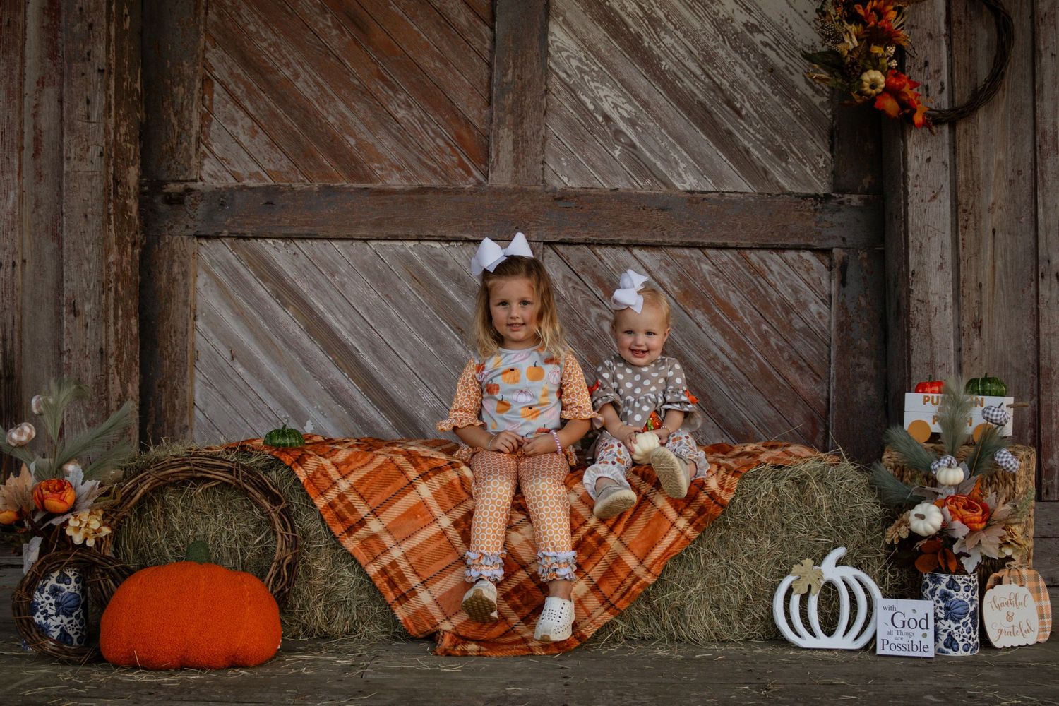 Two young sisters in fall outfits sitting on hay bale with orange plaid blanket and pumpkins.