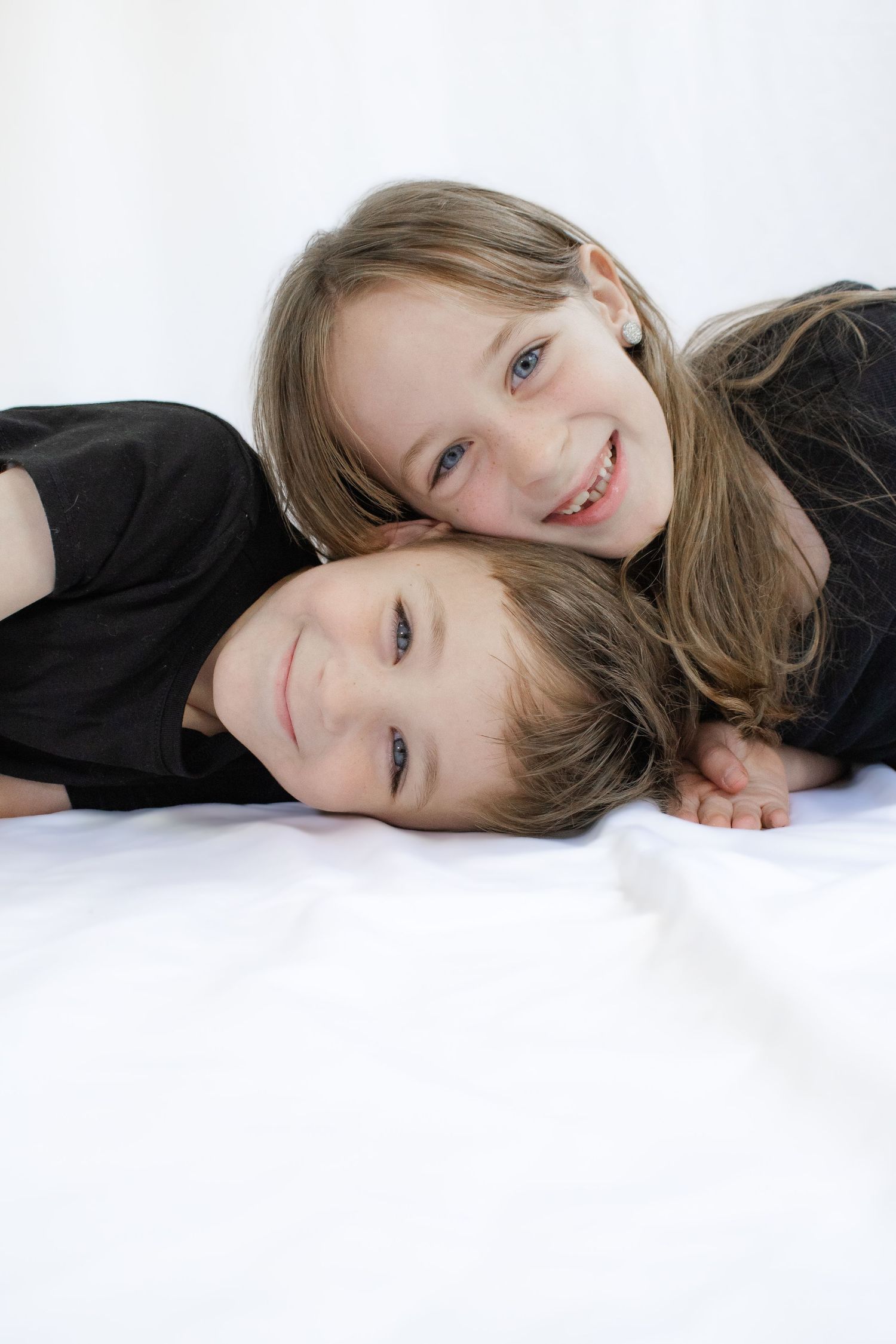 Two smiling children in black shirts lying on a white surface, with the older girl resting her chin on the younger child.