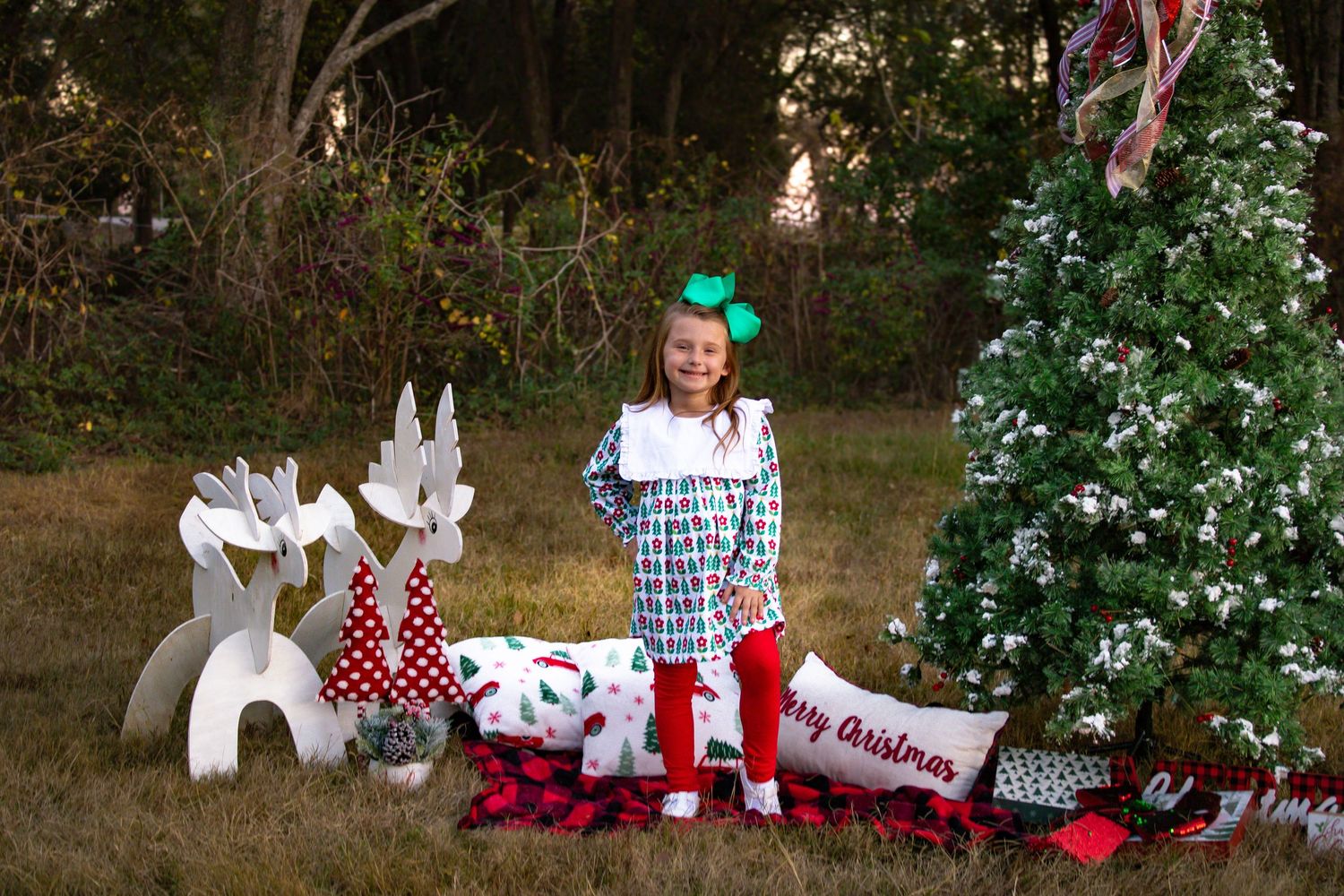 Young girl in plaid dress with green bow standing by Christmas tree and white reindeer props.