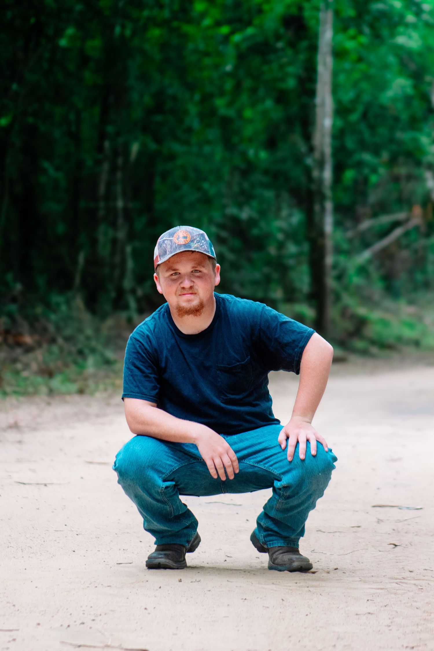 Man in black t-shirt and camo cap crouching down on a dirt road with green trees in background.