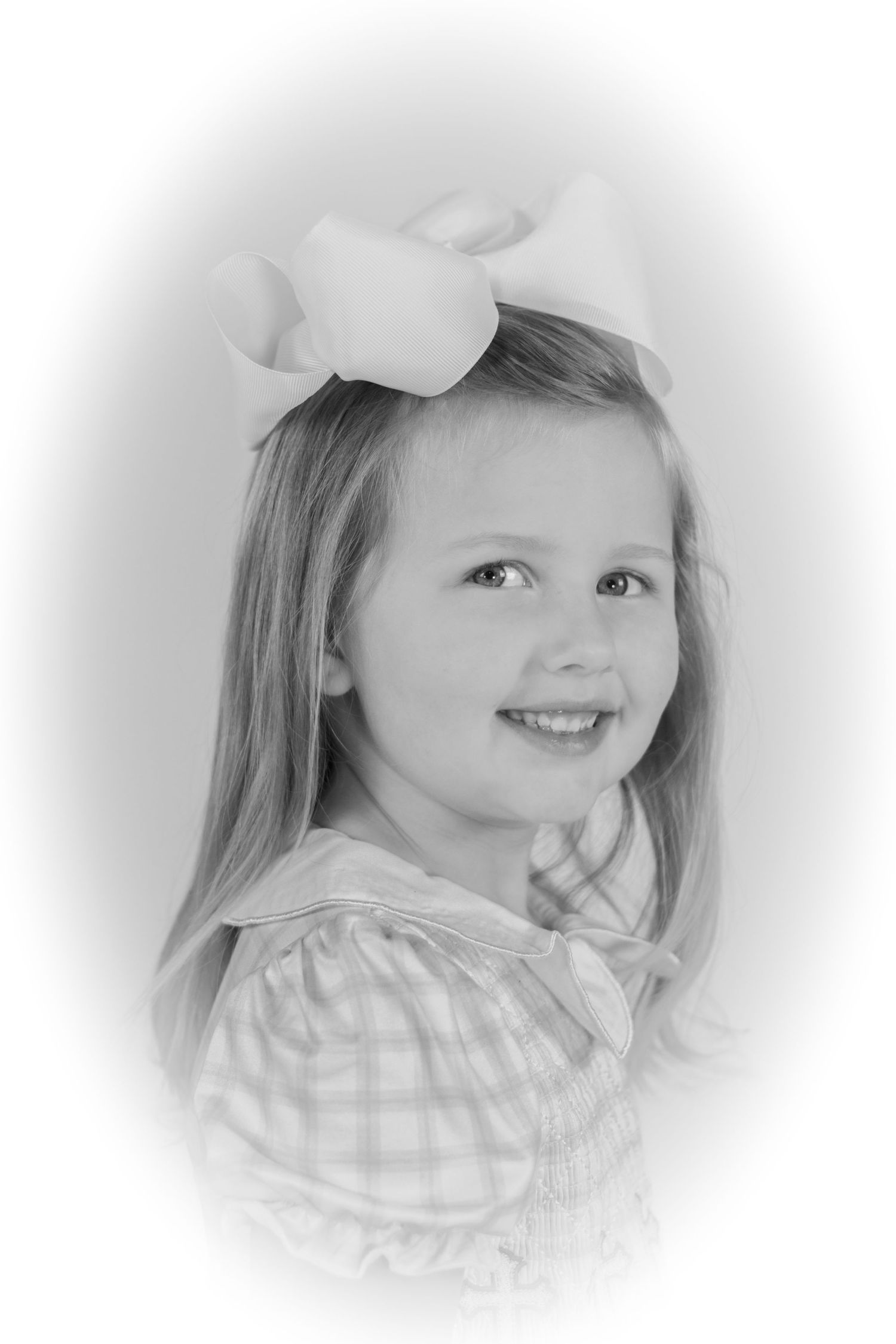 Black and white portrait of a smiling young girl with long hair wearing a large white bow and plaid dress.