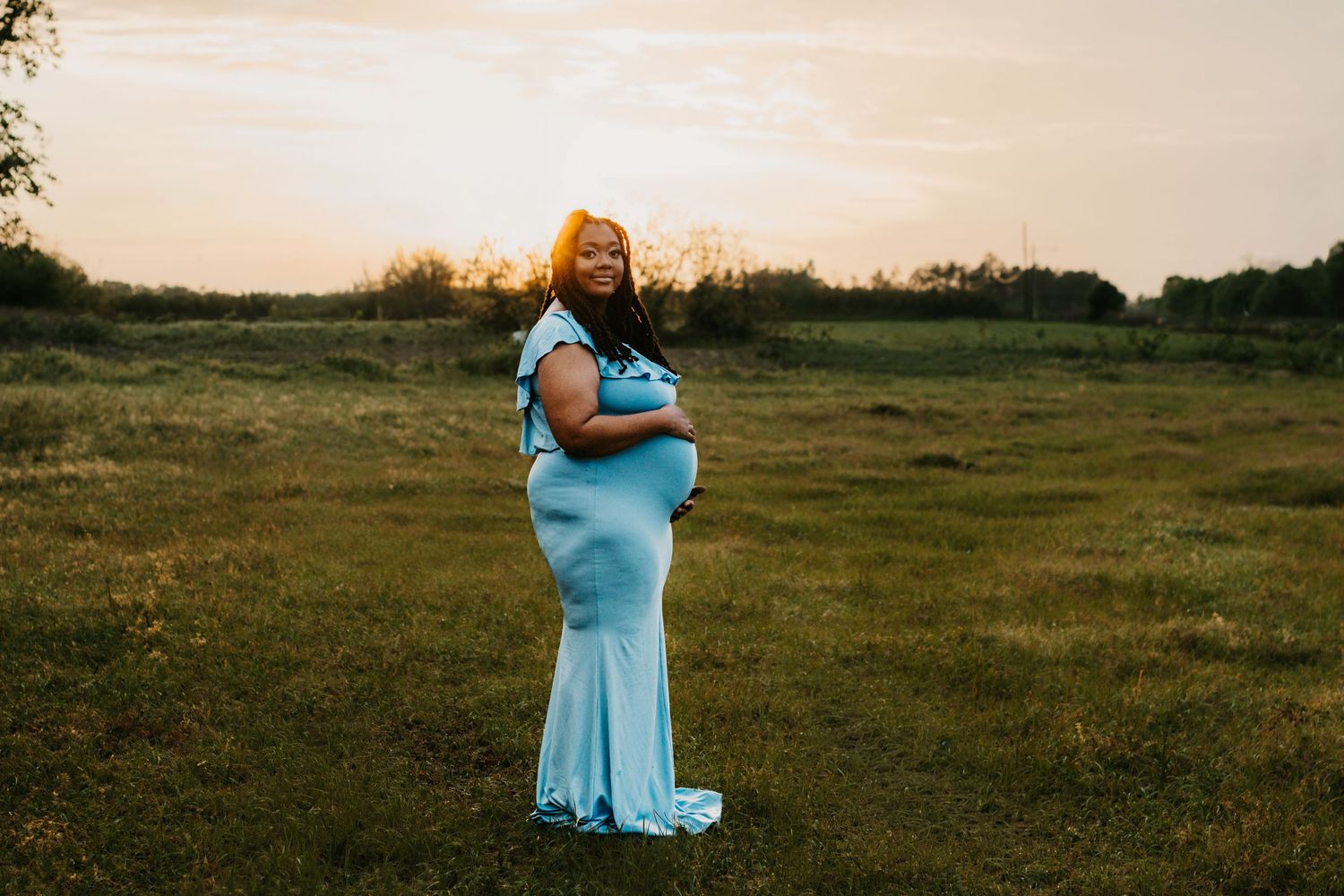 Radiant pregnant woman in light blue maternity gown cradles baby bump in golden sunset meadow.