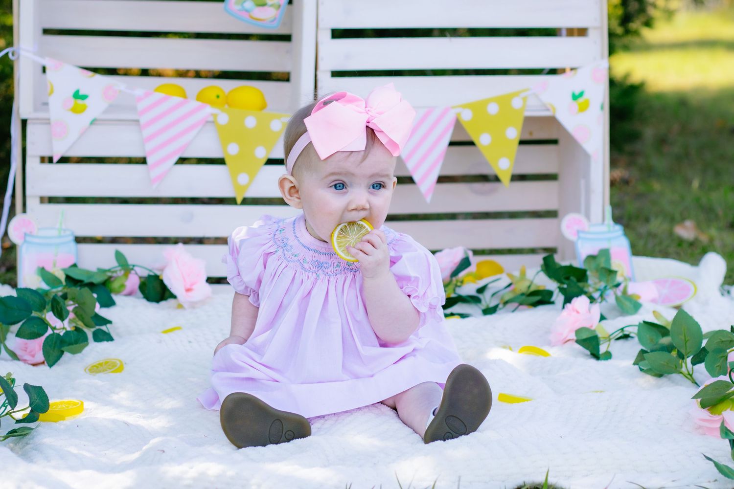 Baby girl in pink dress with bow headband sits on white blanket outdoors near pastel bunting flags.