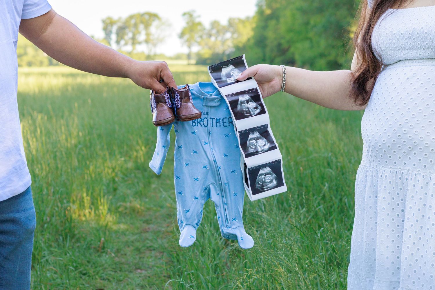 Couple holds baby boy onesie and ultrasound photos in a green field for a gender reveal announcement.