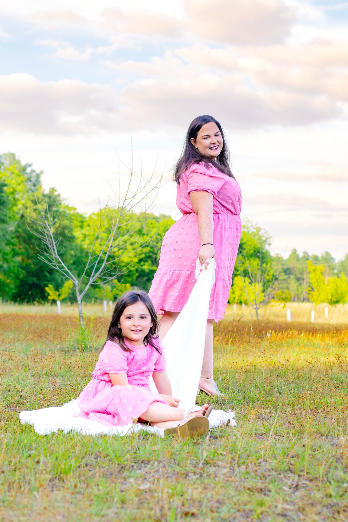 Pregnant woman and young daughter in matching pink dresses pose outdoors in a green field under a cloudy sky.