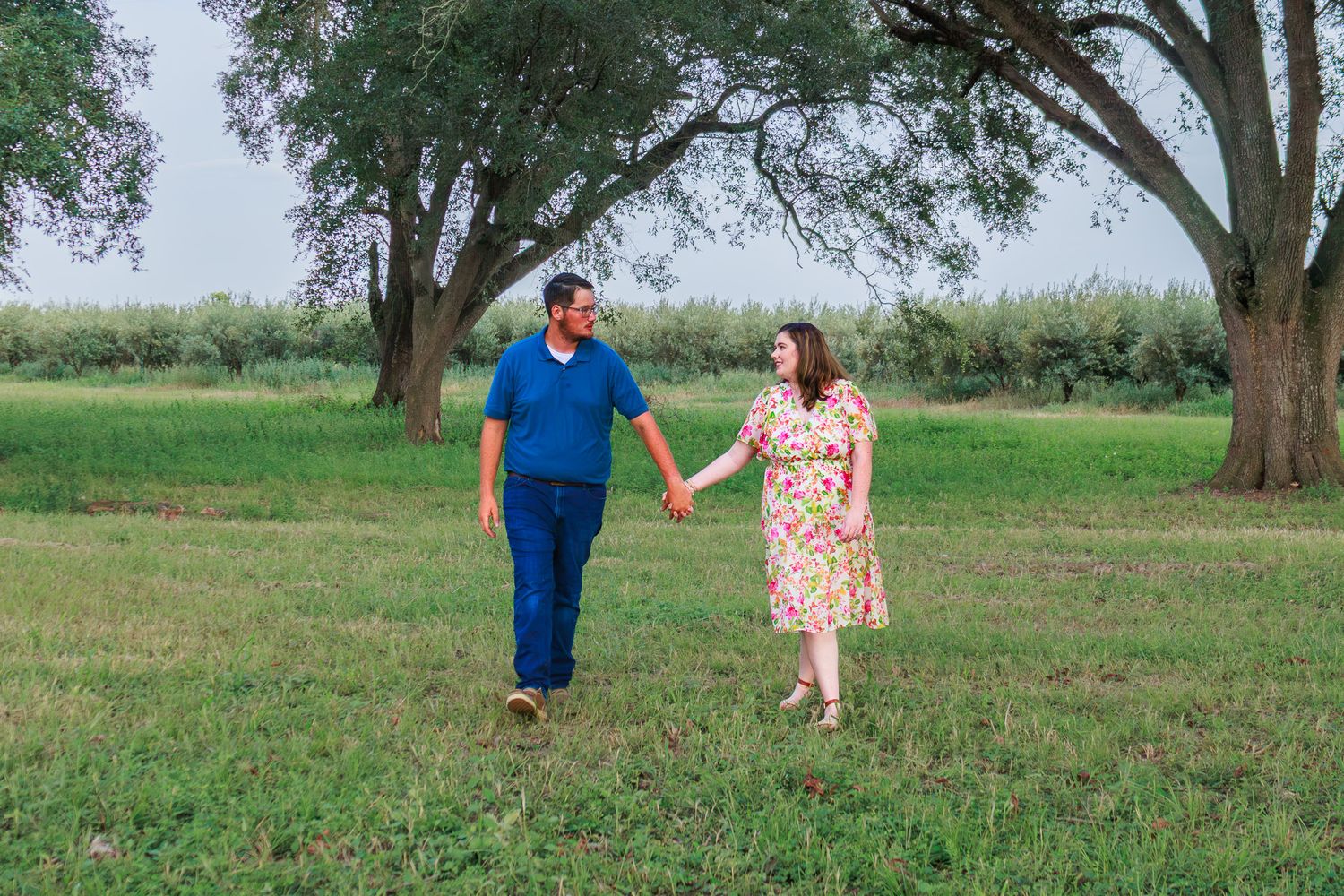 Couple holding hands walking through a lush green park with large trees on a cloudy day.