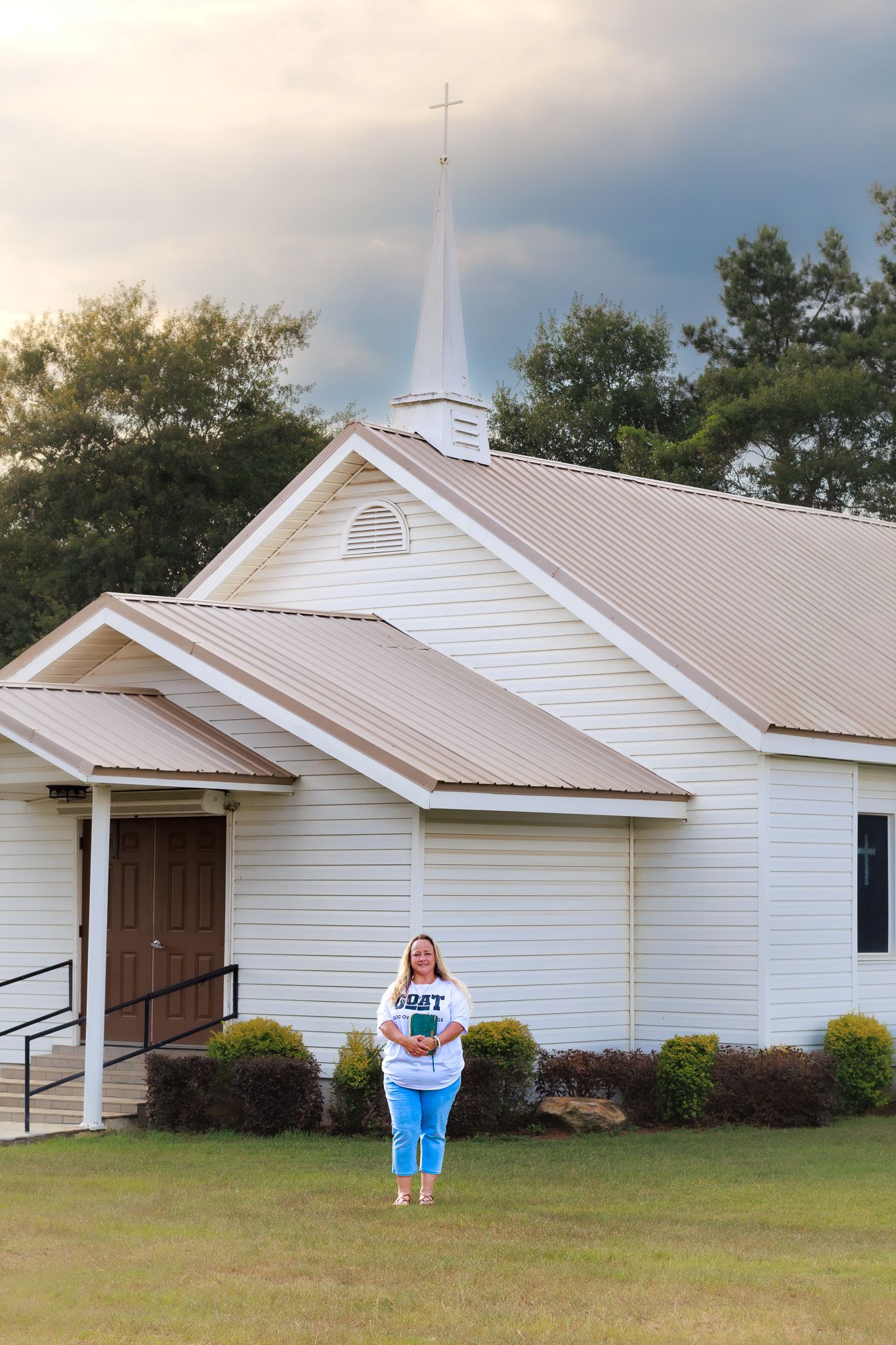 Woman holding Bible standing outside white rural church with steeple under dramatic cloudy sky.