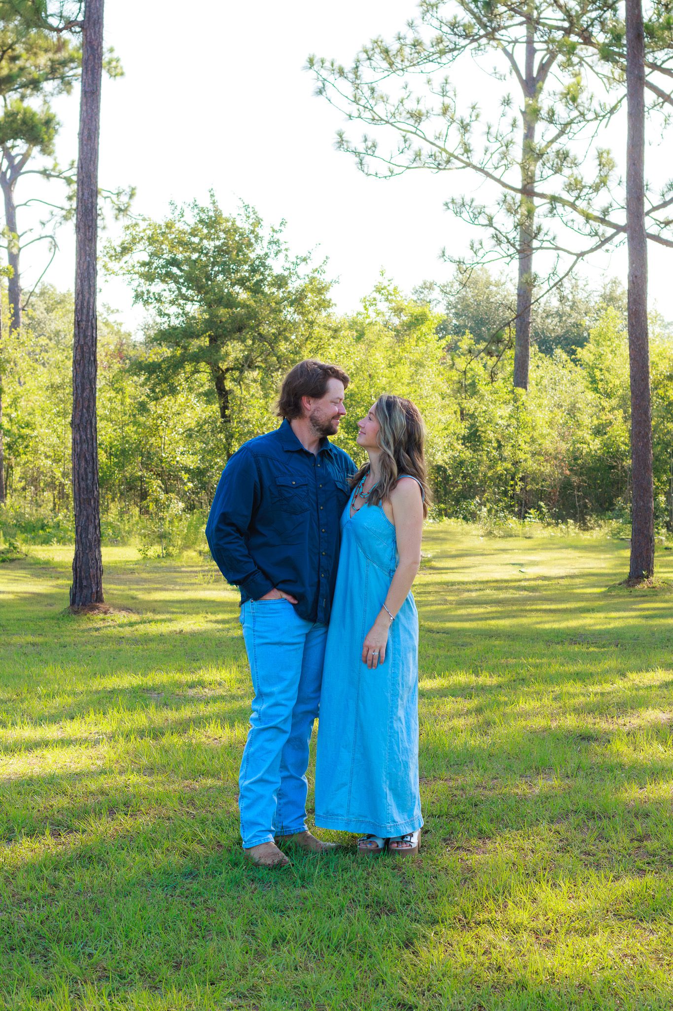 Couple in coordinating blue outfits share a romantic moment standing on green grass surrounded by tall pine trees.