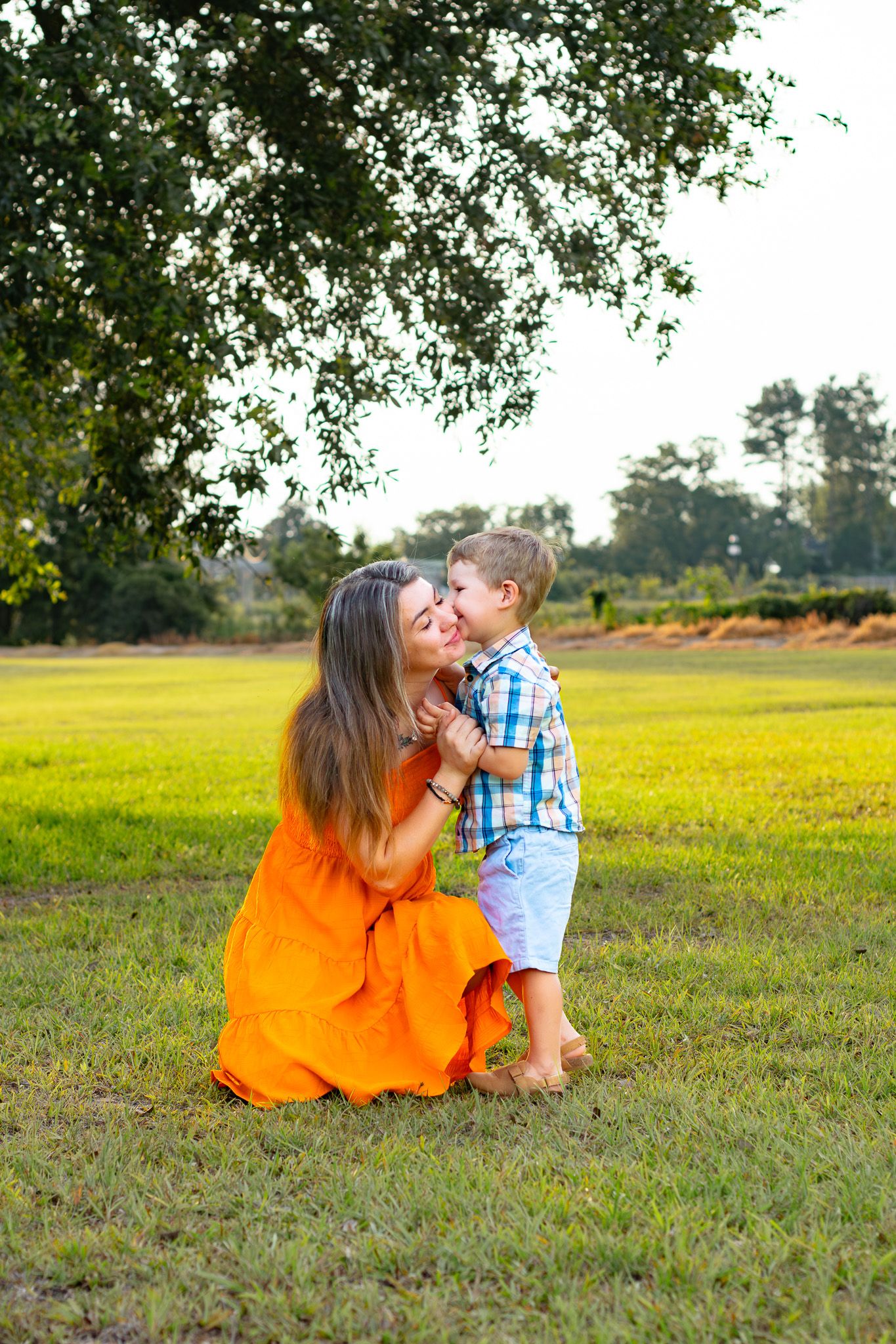 A mother in an orange dress kneels on green grass, sharing a tender kiss with her young son outdoors.