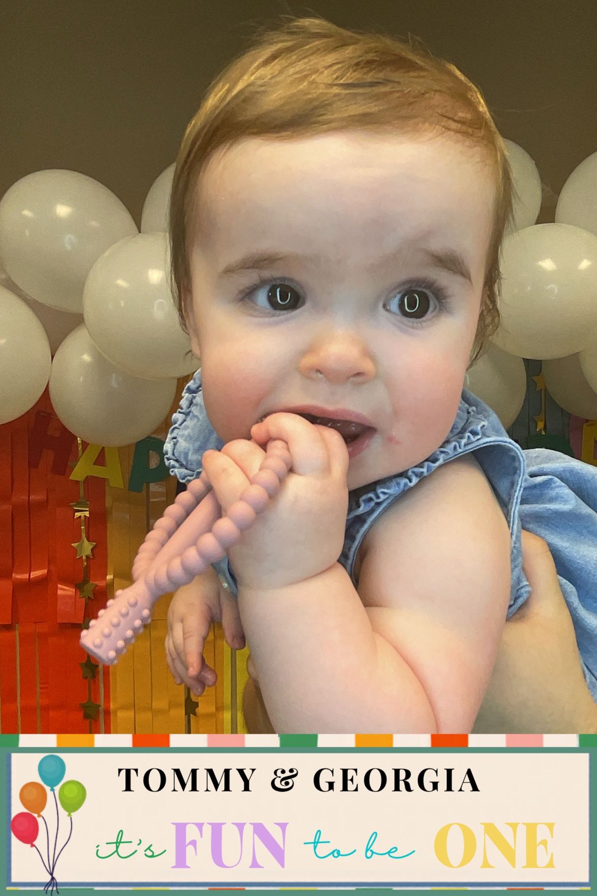 Baby girl chewing on pink teether toy at Tommy & Georgia's first birthday party with balloon backdrop.