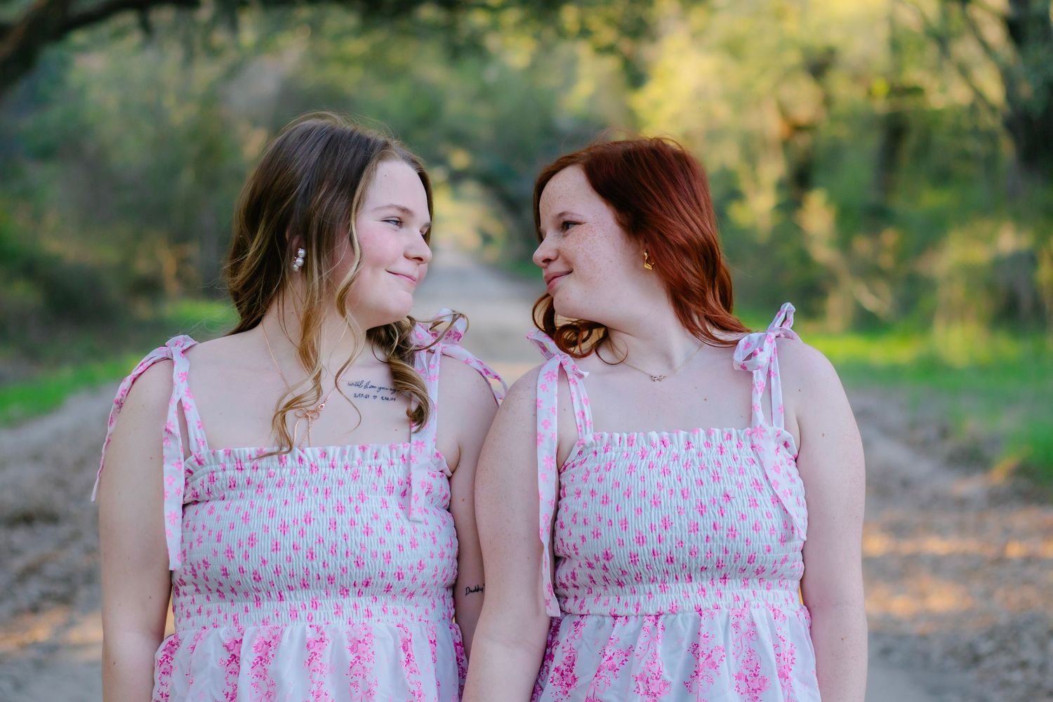 Two smiling young women in matching pink floral sundresses gaze at each other outdoors.