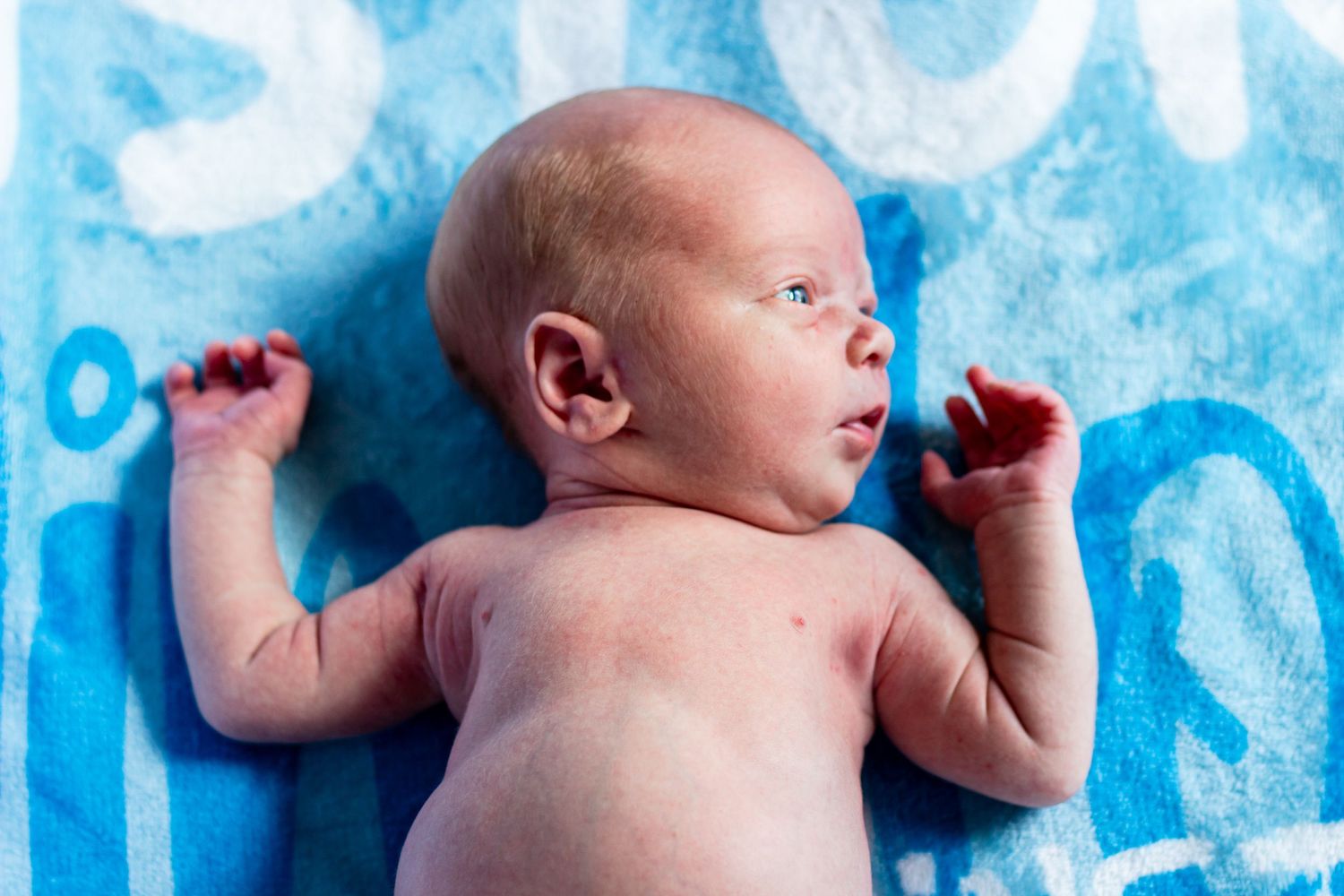 Newborn baby lying on a blue blanket with text, eyes open, tiny fingers spread wide.