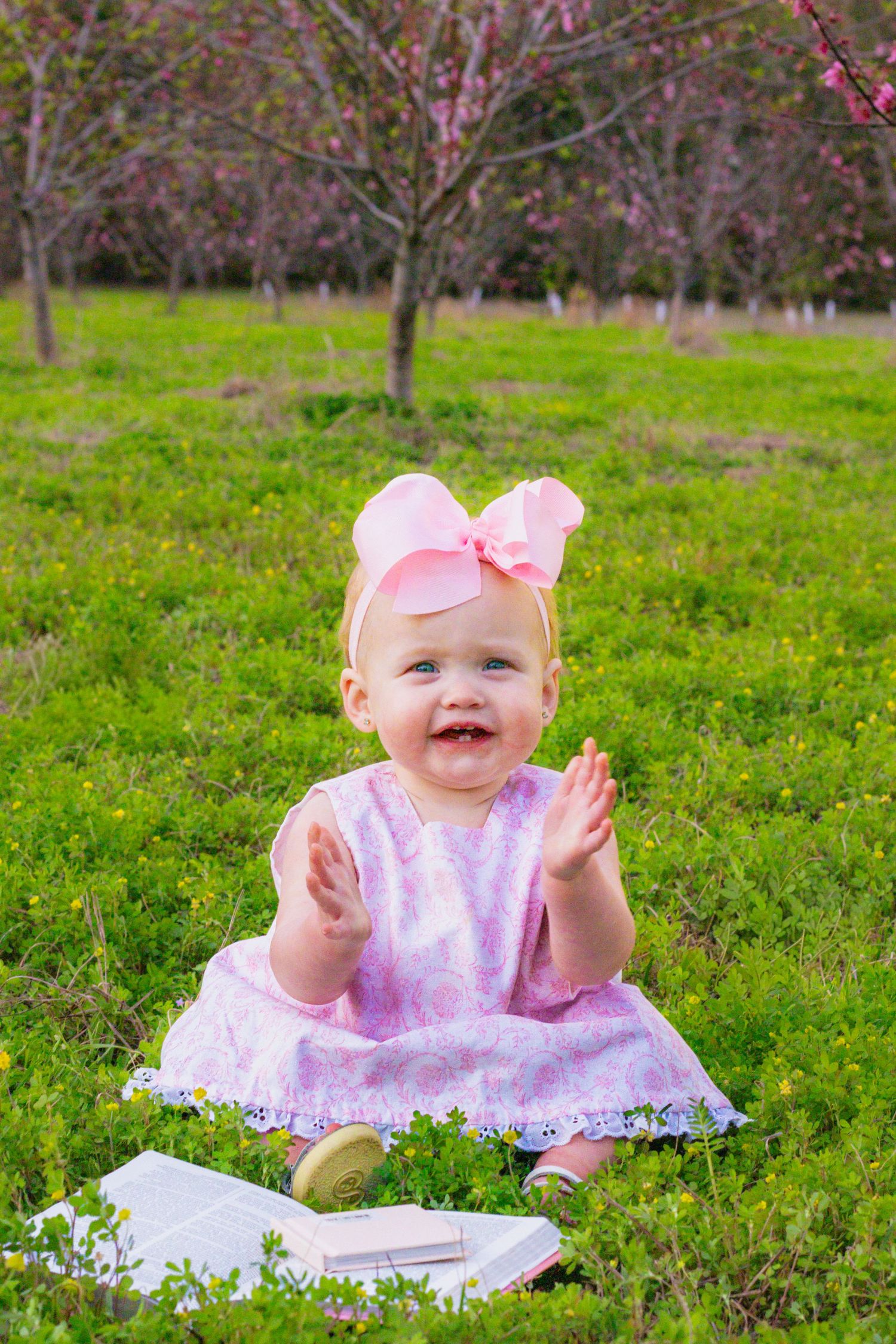 Happy baby girl in pink dress and bow clapping hands while sitting on green grass outdoors.