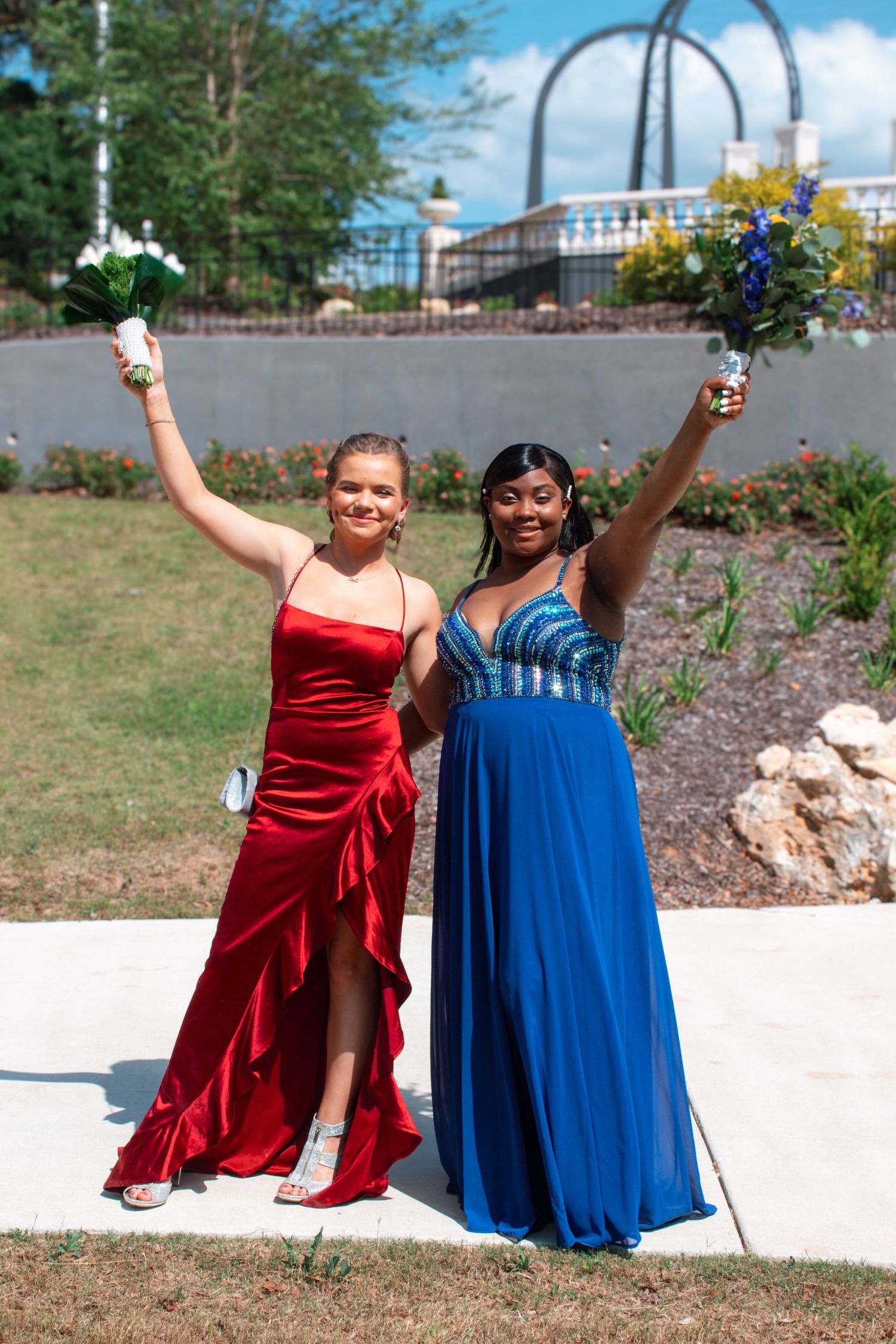 Two young women in red and blue formal prom dresses raising bouquets outdoors on a sunny day.