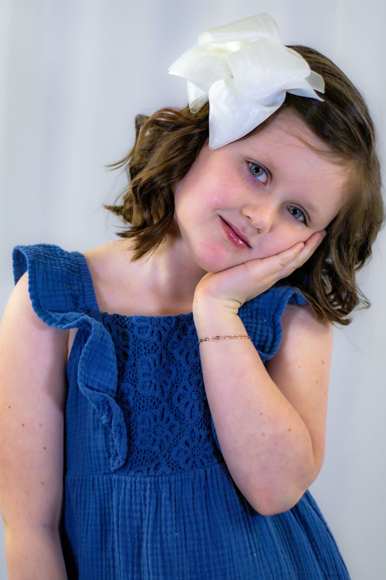 Sweet girl in blue dress with white bow resting cheek on hand dreamily.