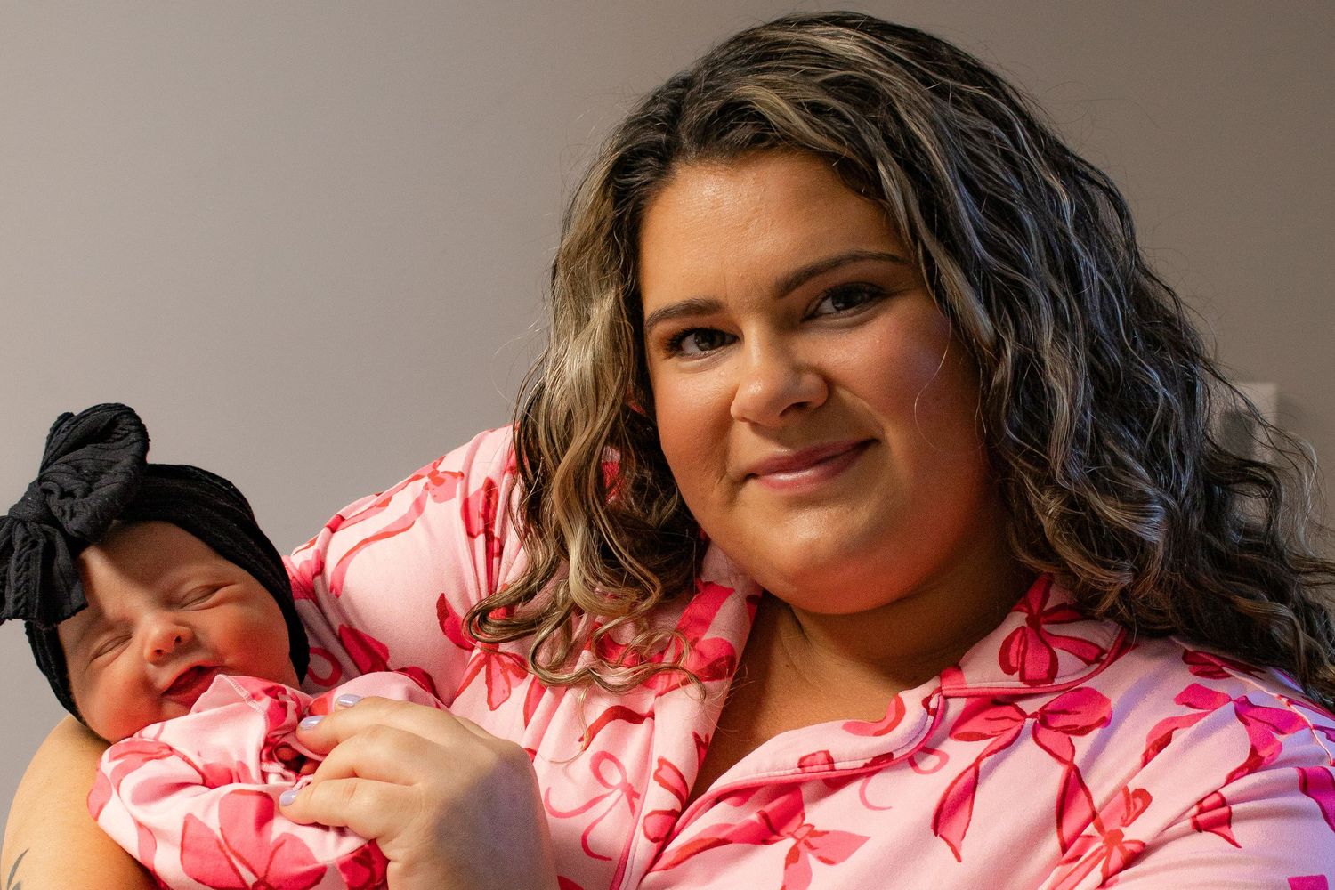 A smiling young mother in a pink floral gown holds her newborn baby with a black bow headband.