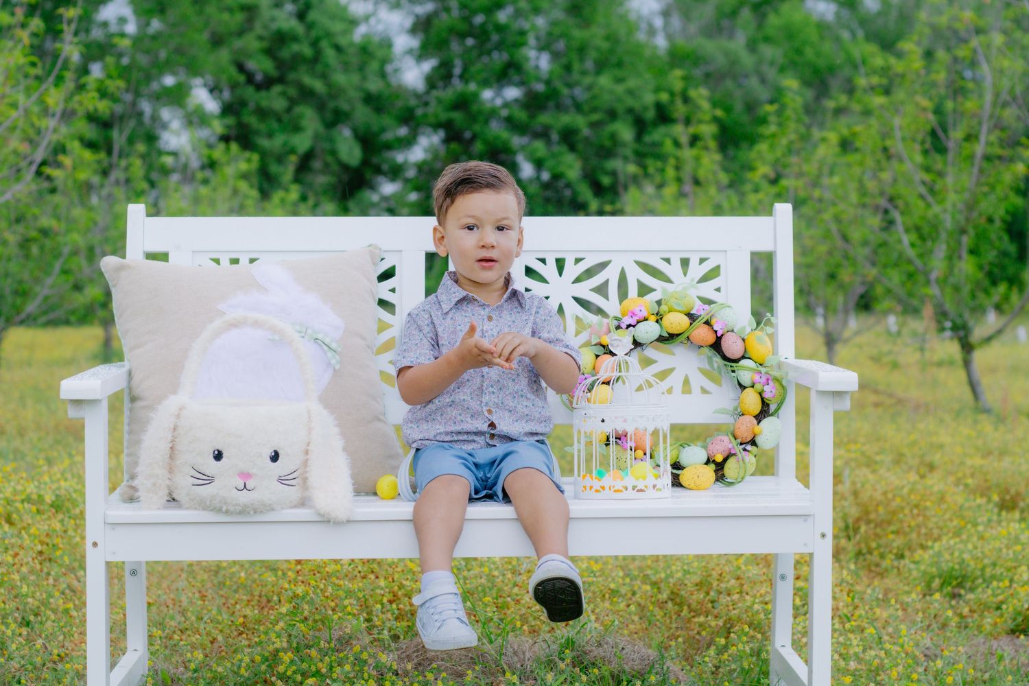 Young boy sitting on white garden bench with stuffed Easter bunny and colorful wreath outdoors.