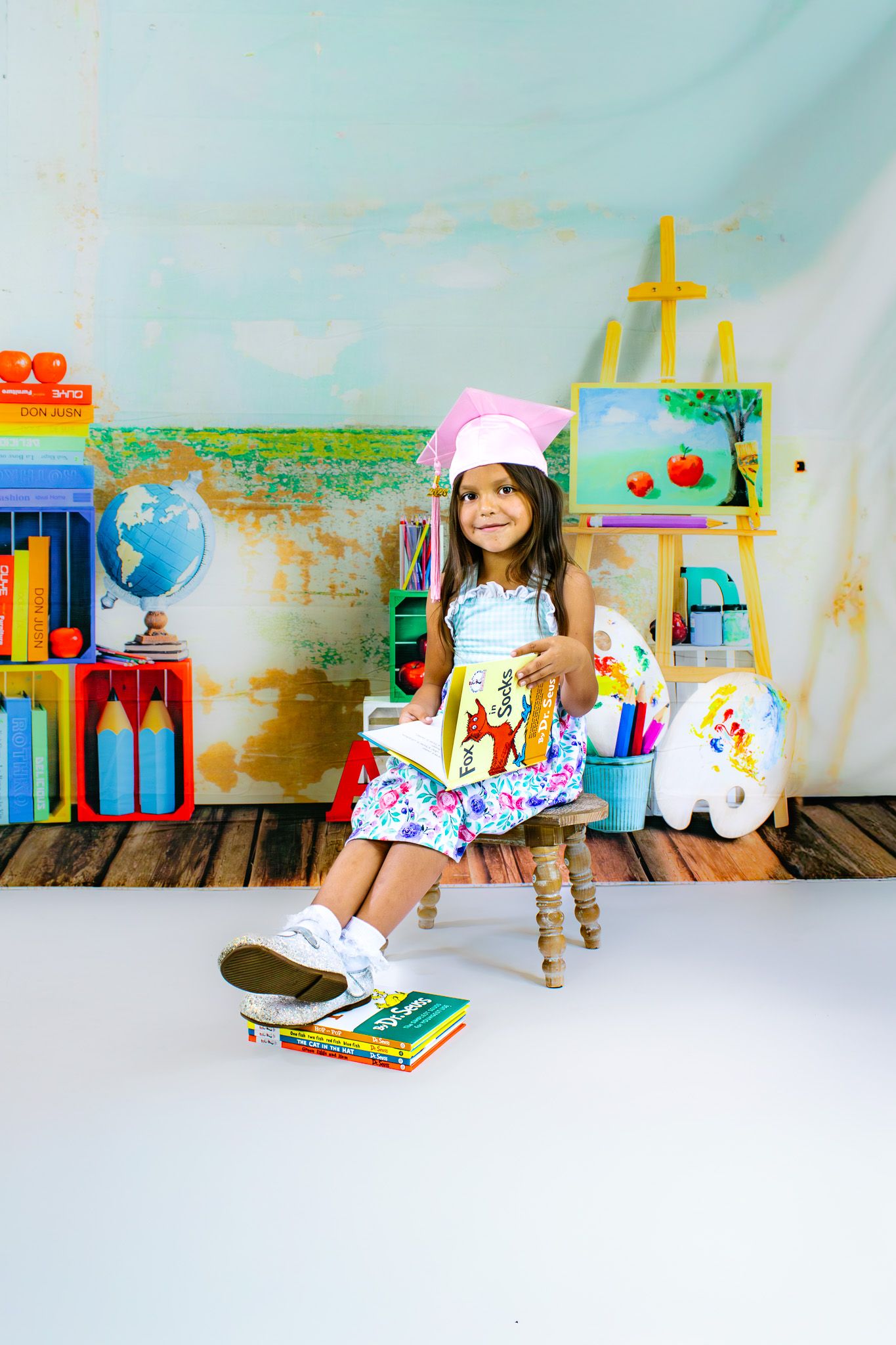 Young girl in floral dress and pink graduation cap sits on chair holding book, surrounded by colorful school supplies.