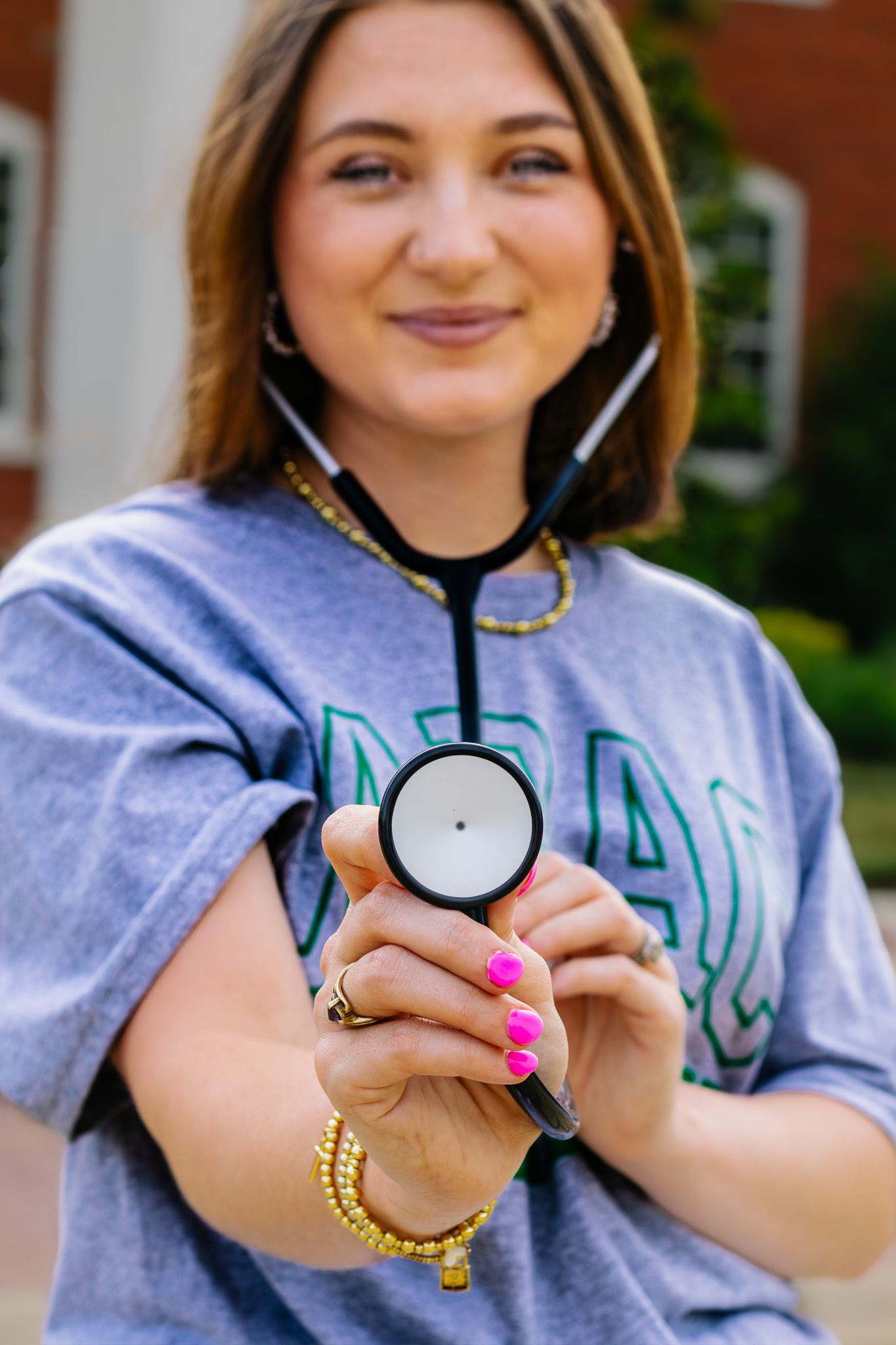 Smiling nursing student holds stethoscope toward camera wearing gray NRAC college t-shirt outdoors.