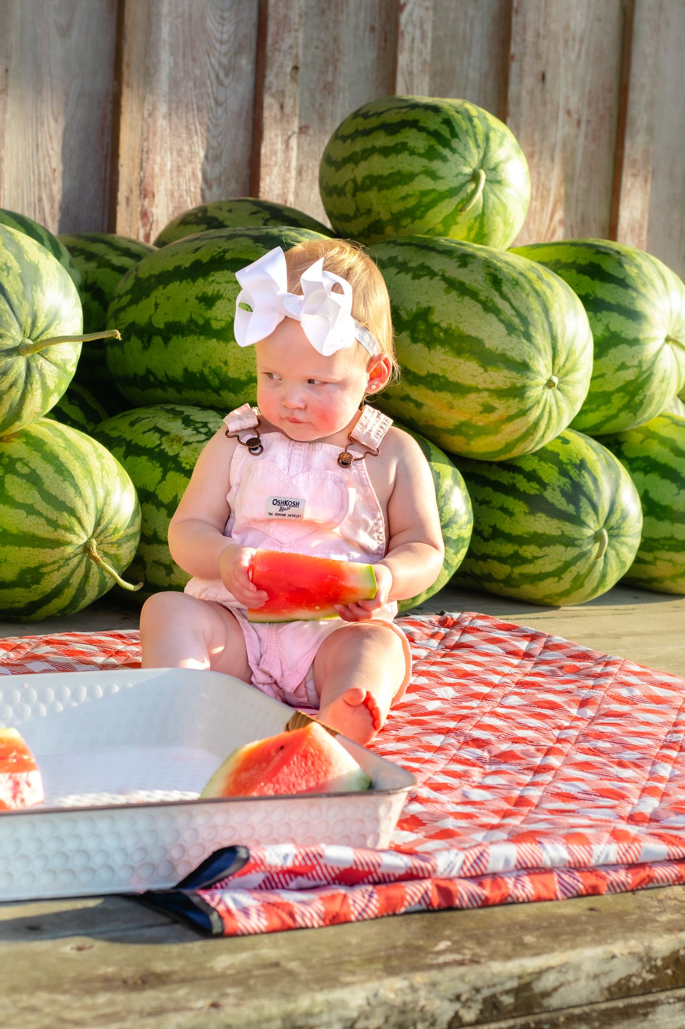Baby girl in pink OshKosh overalls examines a watermelon slice while sitting on red checkered blanket by watermelons.