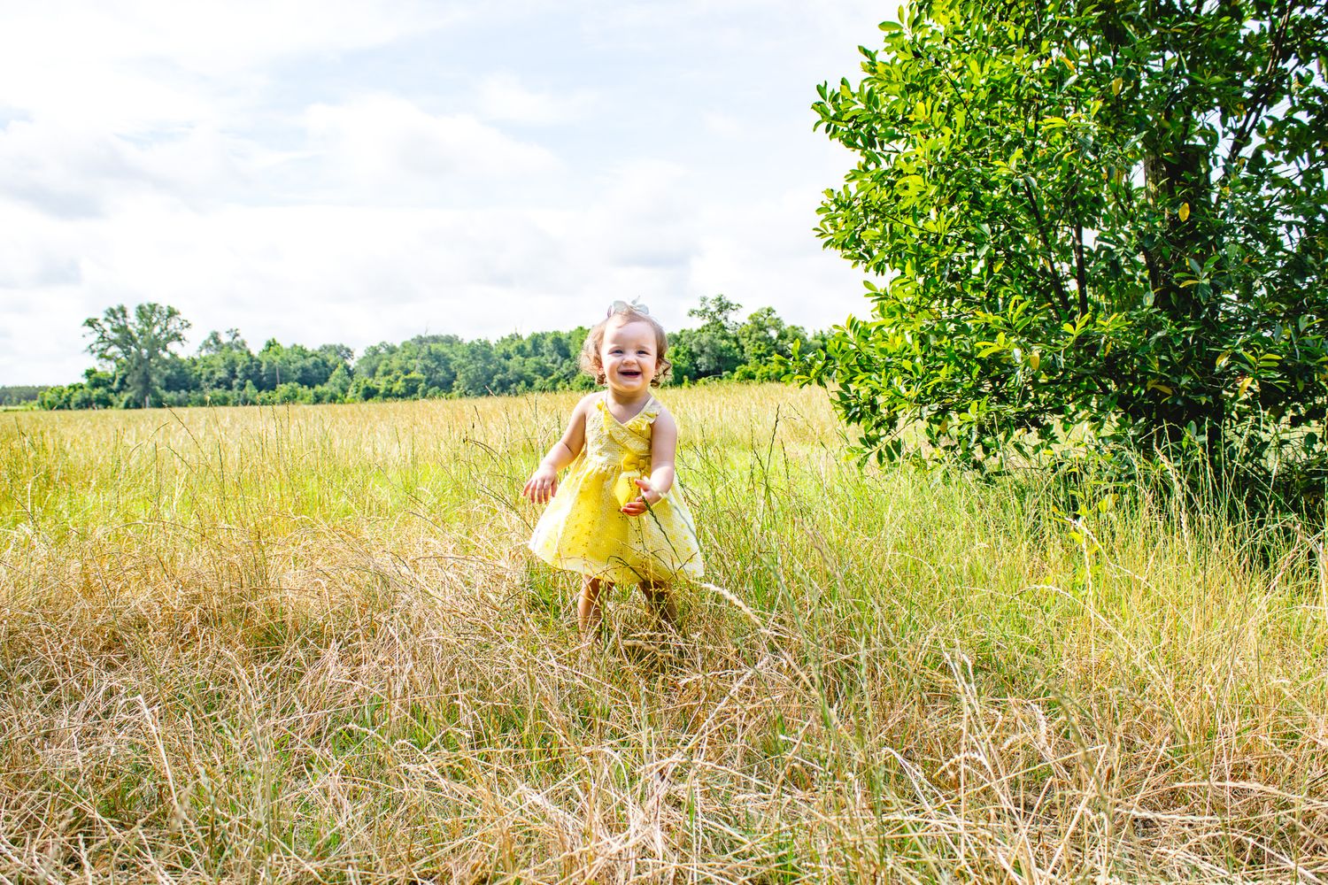 A joyful toddler in a yellow dress runs through a golden meadow with green trees and cloudy sky behind her.