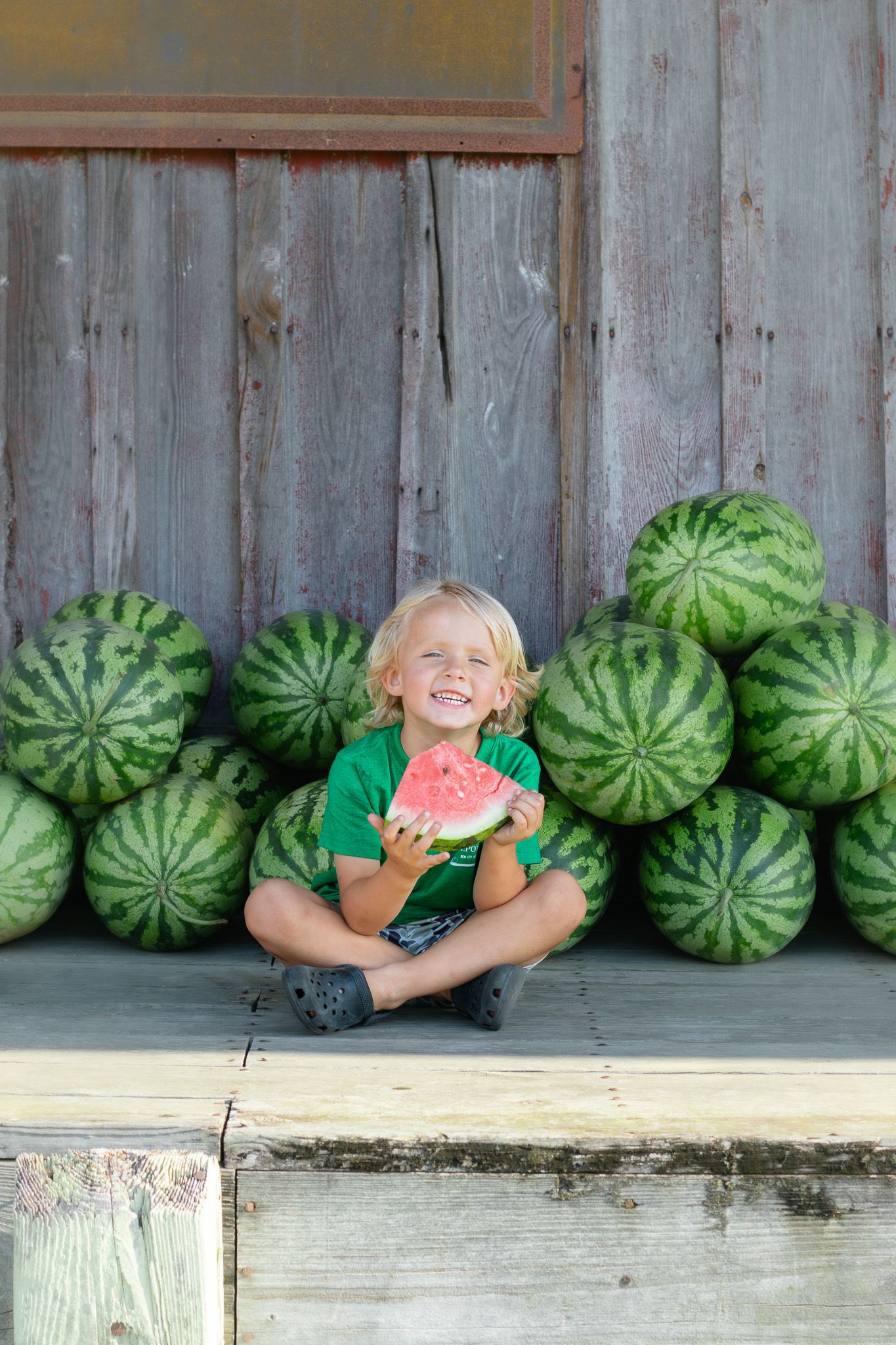 Smiling blonde child holding watermelon slice while sitting cross-legged in front of stacked watermelons against weathered barn wall.