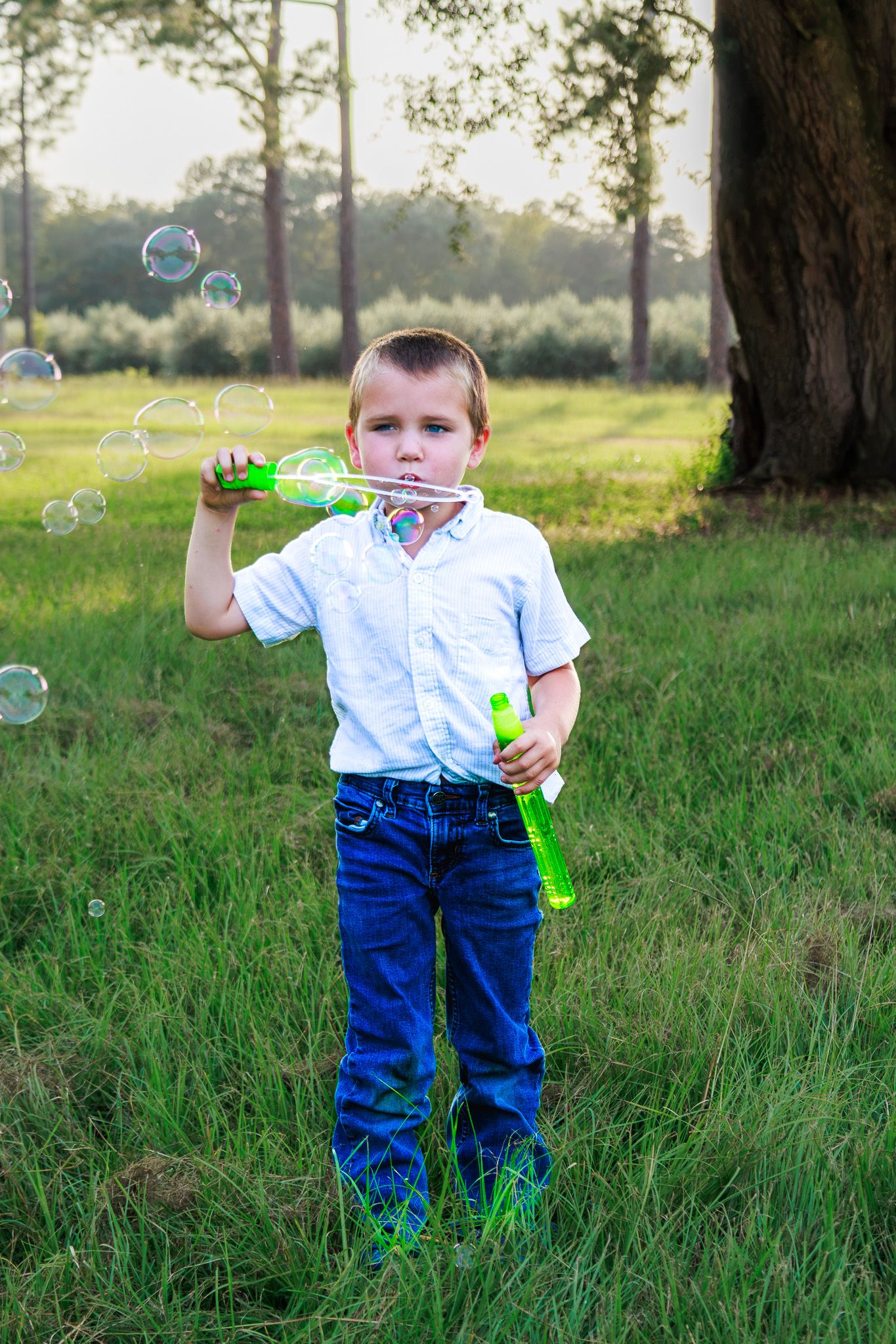 Young boy in white shirt and jeans blowing soap bubbles with a green wand in a sunny green park.