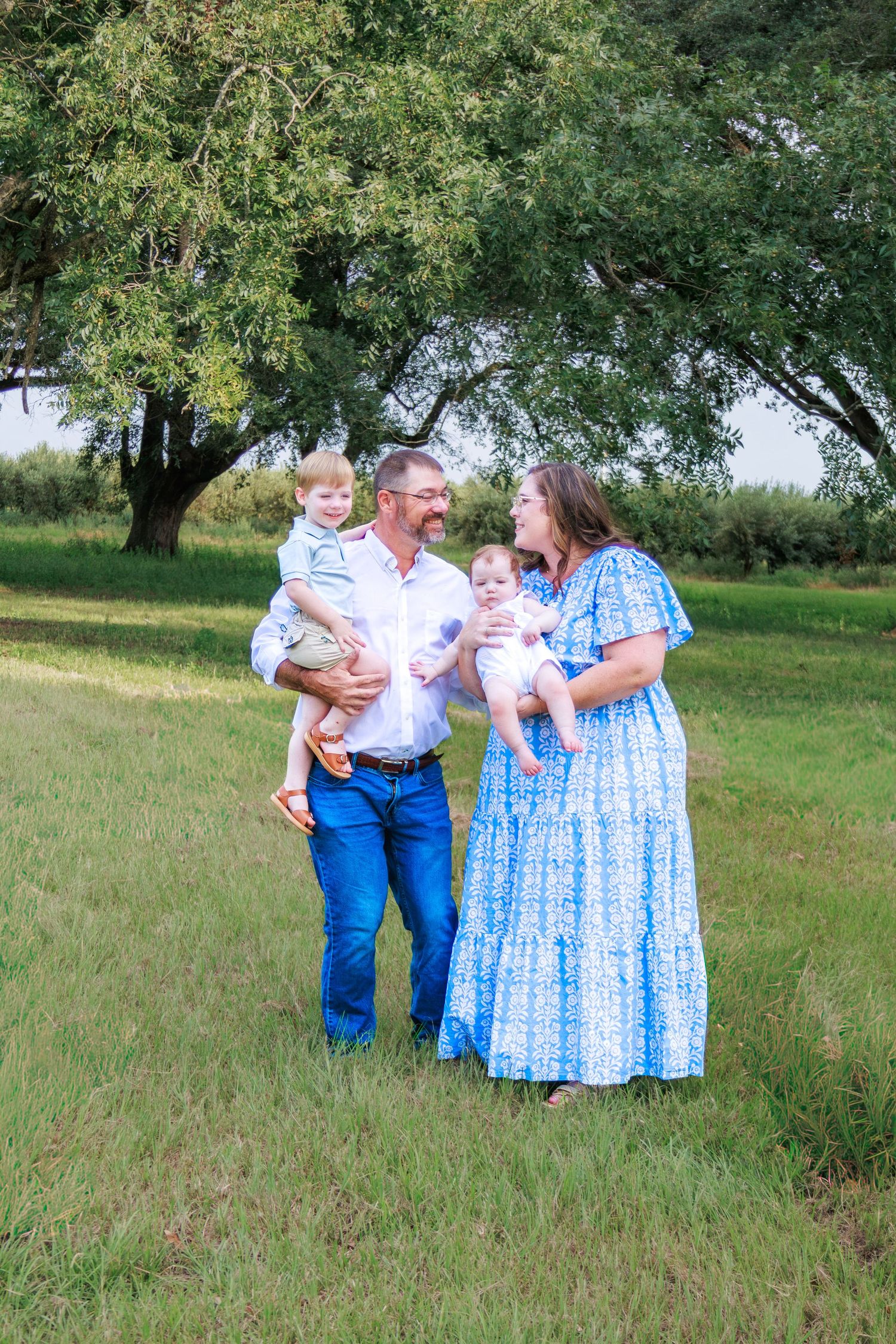 A happy family of four poses outdoors, parents holding two young children in a lush green field with trees.