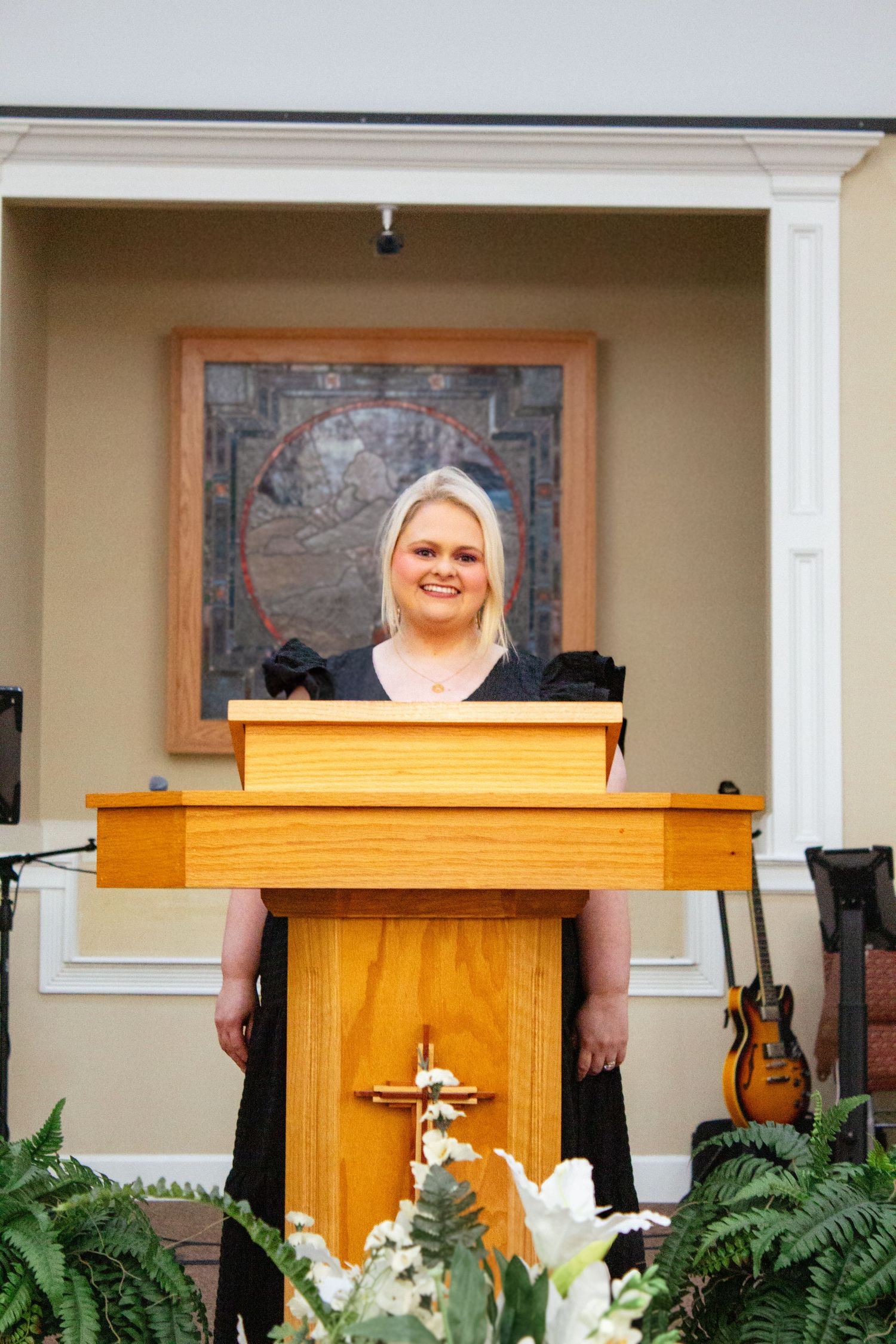 Blonde woman in black dress smiling at church pulpit with cross and floral arrangements.