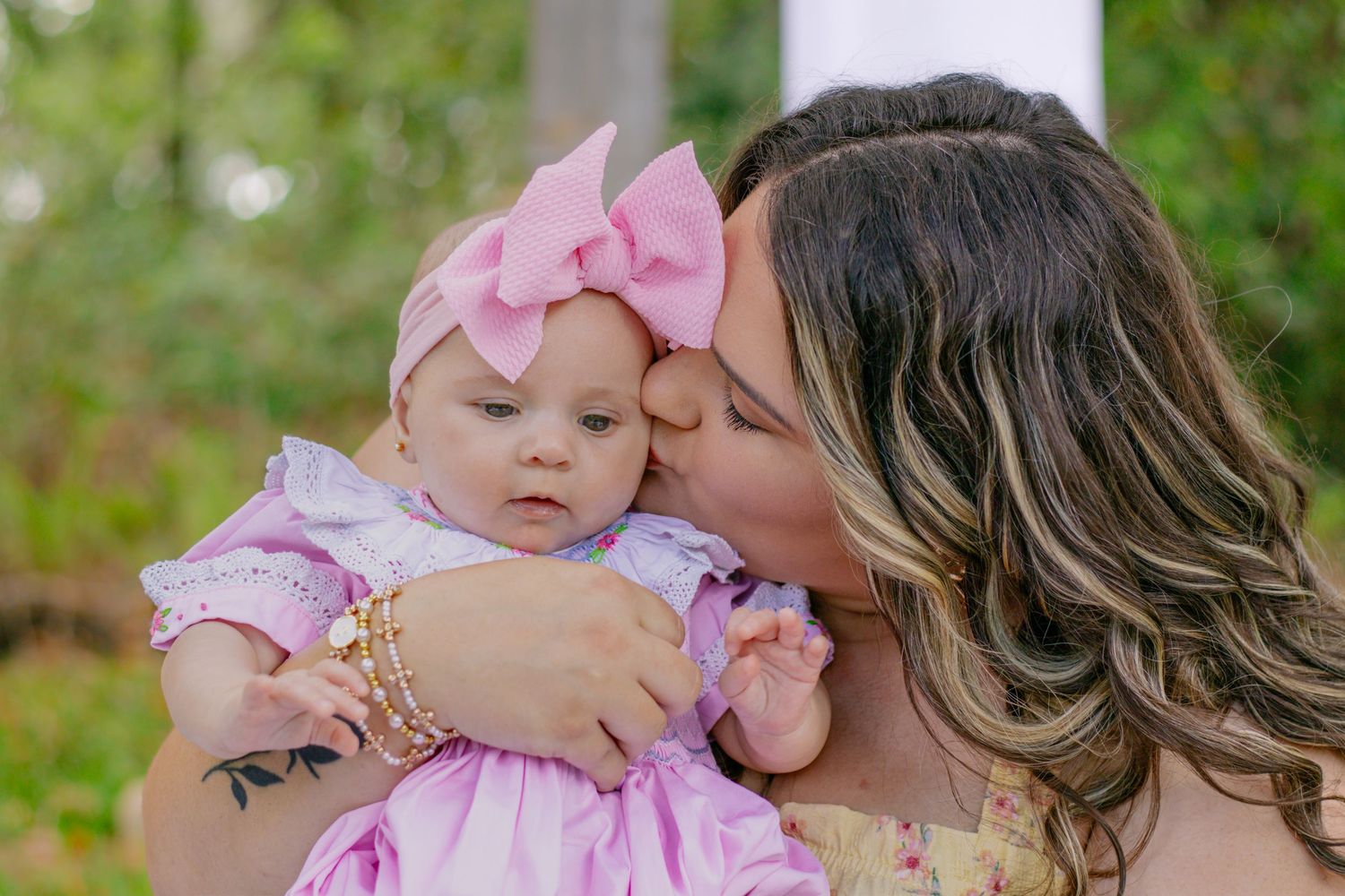 Mother kissing her baby girl dressed in pink smocked outfit with large pink bow headband outdoors.