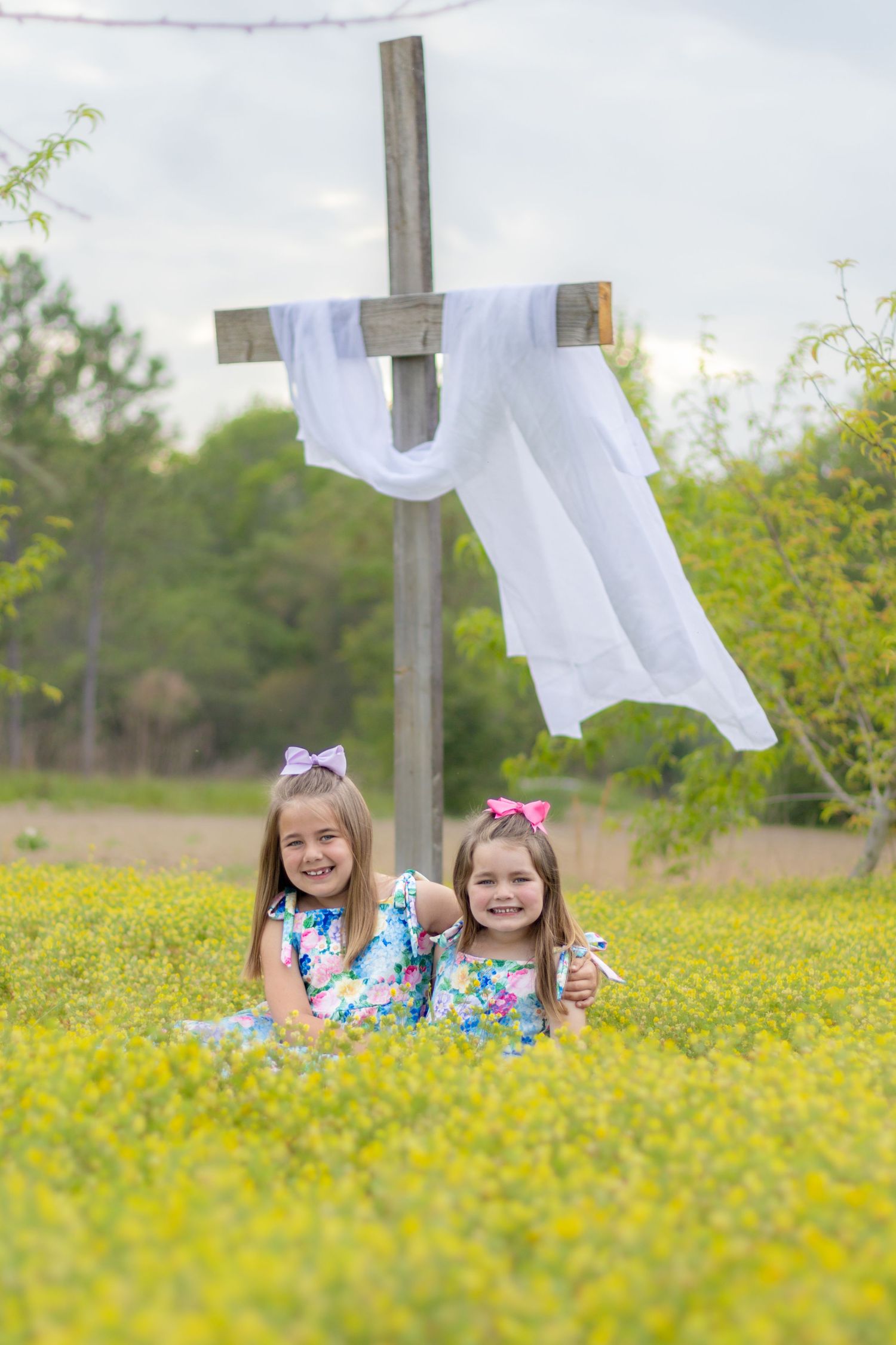 Two little girls in floral dresses sitting together in yellow wildflower field in front of a white-draped wooden cross.