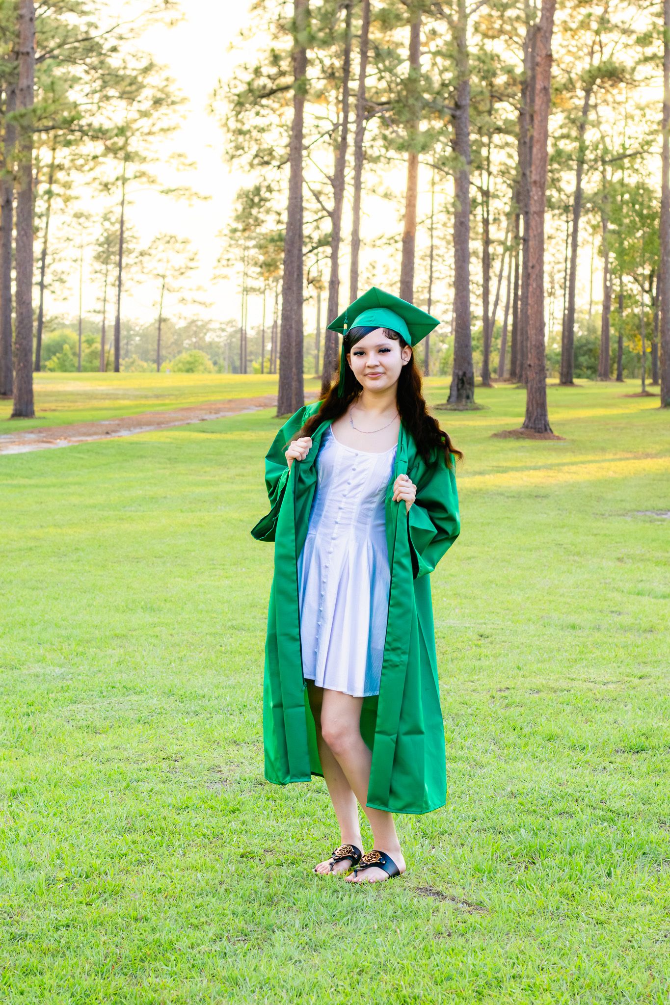 Young woman in green graduation cap and gown posing on grass with tall pine trees in background.