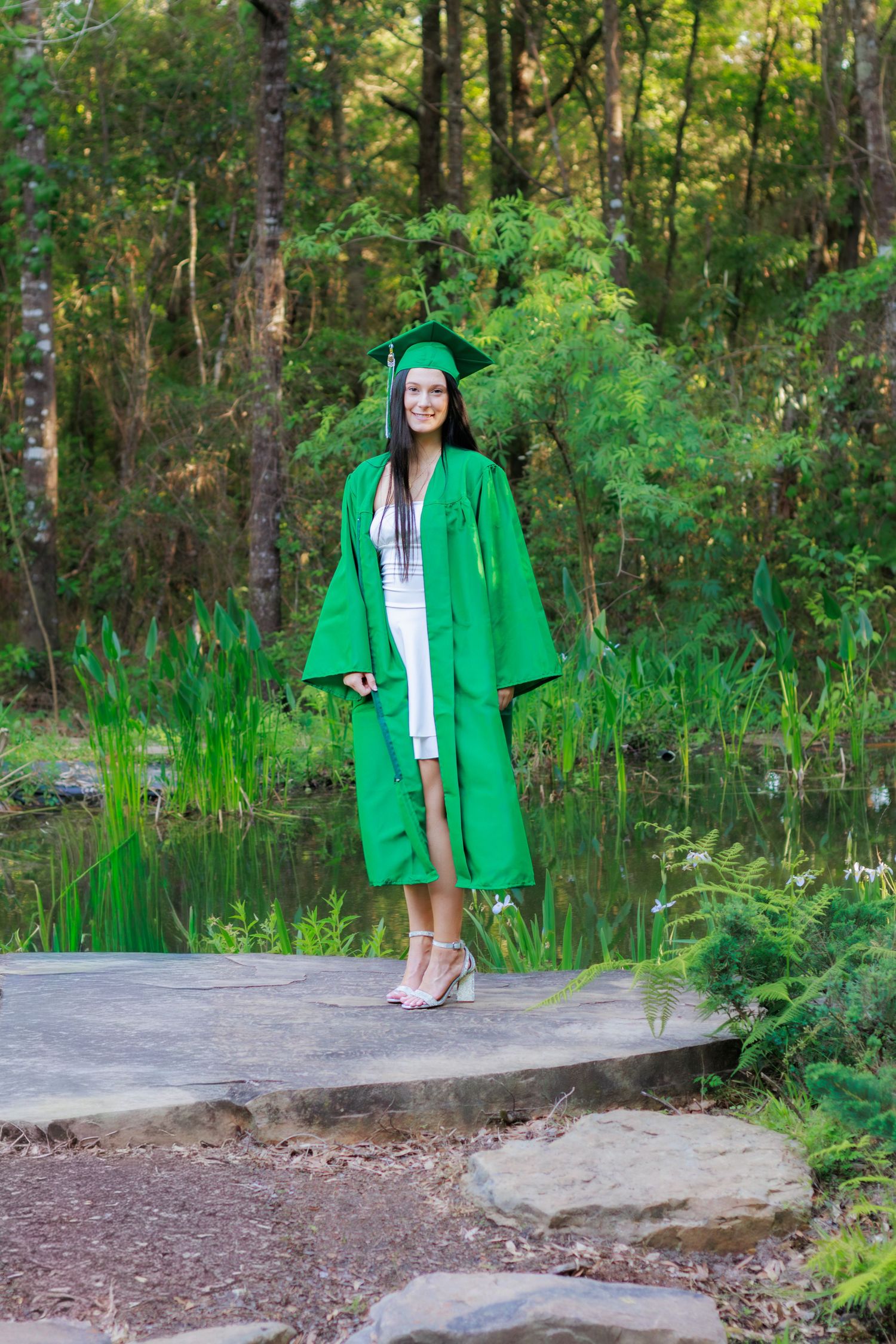 Smiling graduate in green cap and gown stands on stone path beside a pond surrounded by lush greenery.