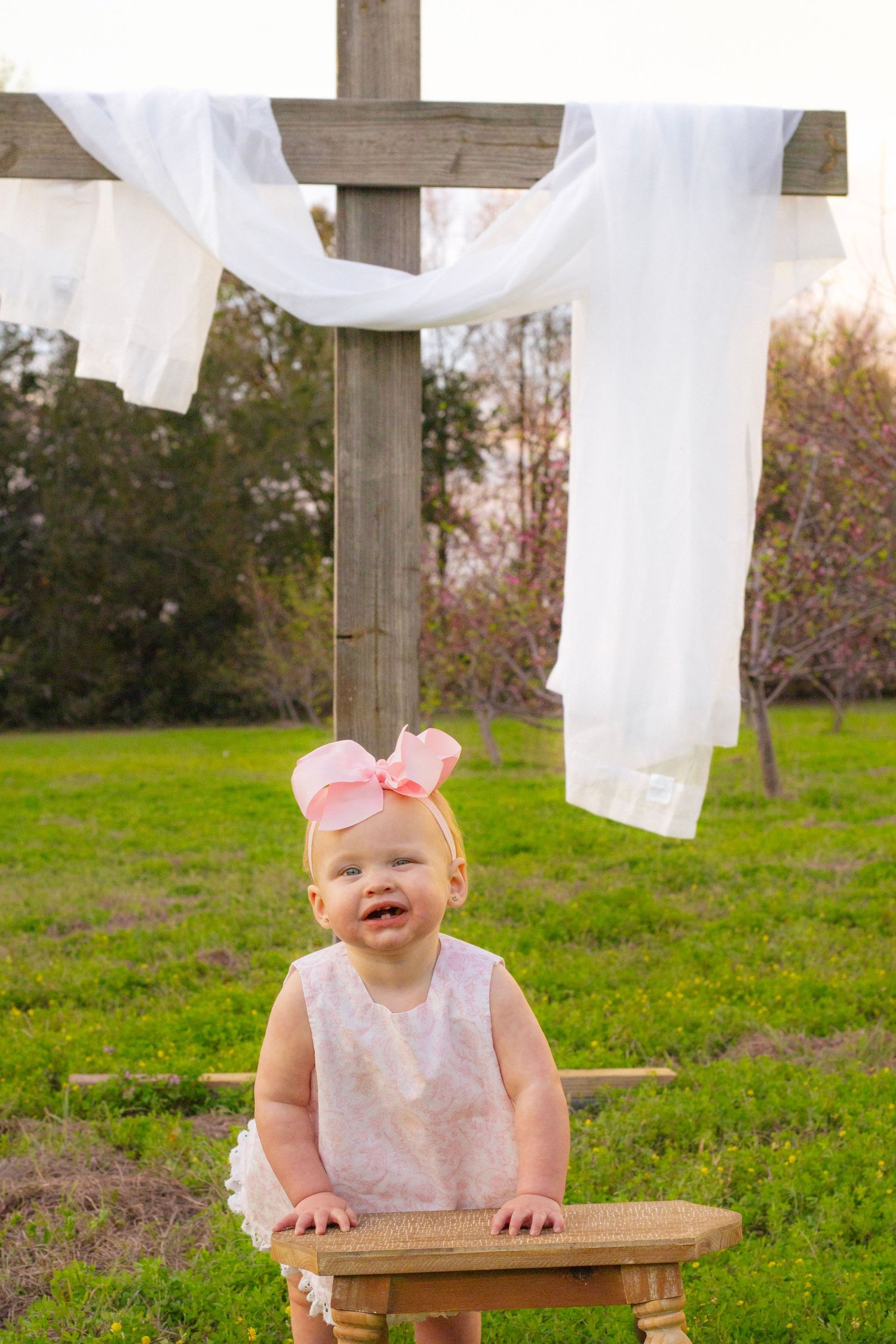 Smiling baby girl in white lace dress leans on wooden stool in front of a rustic cross outdoors.