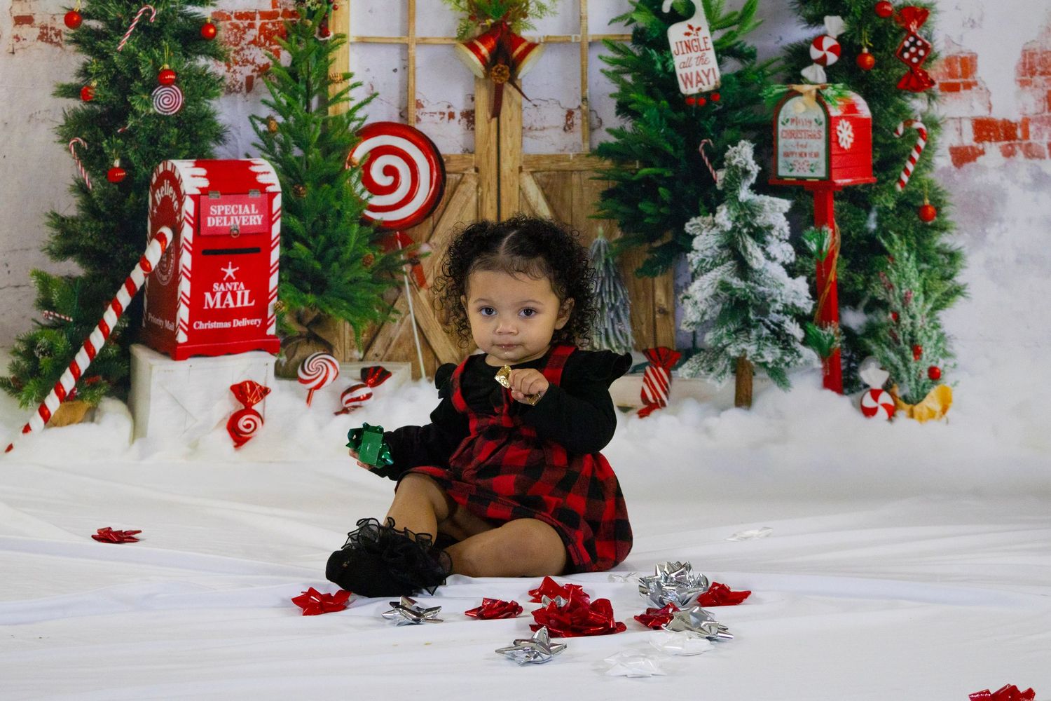 Toddler girl in red plaid dress sits among Christmas decorations during festive holiday photo session.