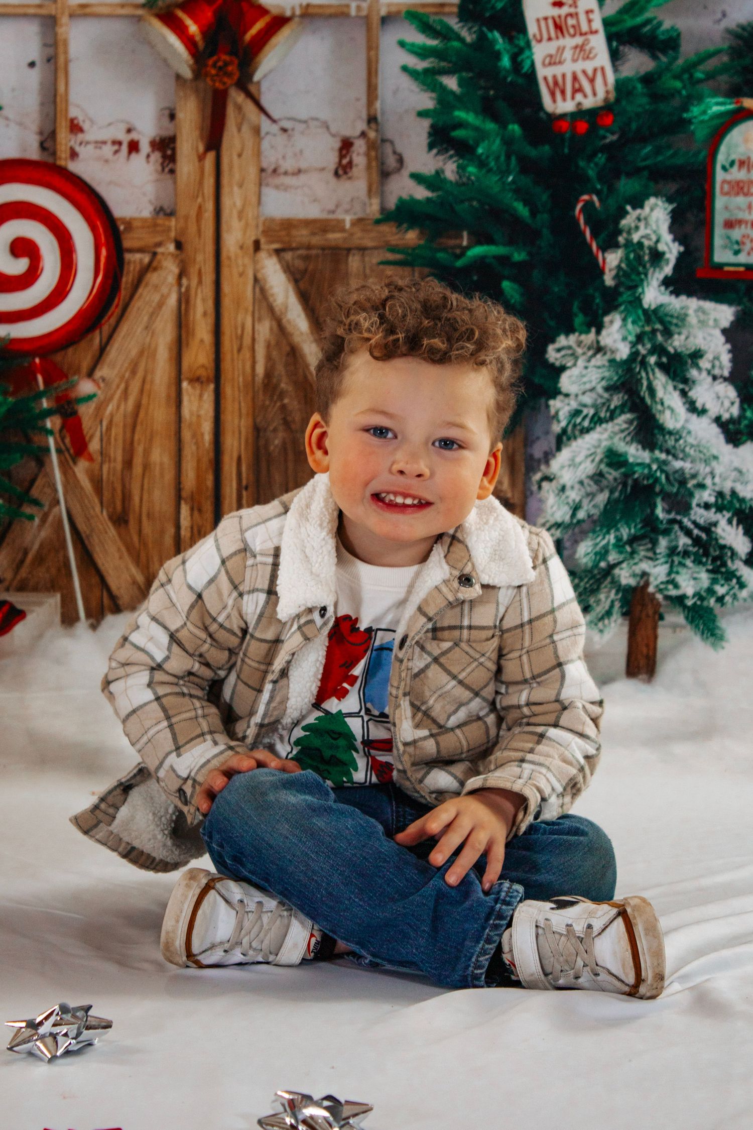 A curly-haired boy in a plaid jacket sits cross-legged on white snow surrounded by Christmas decorations.