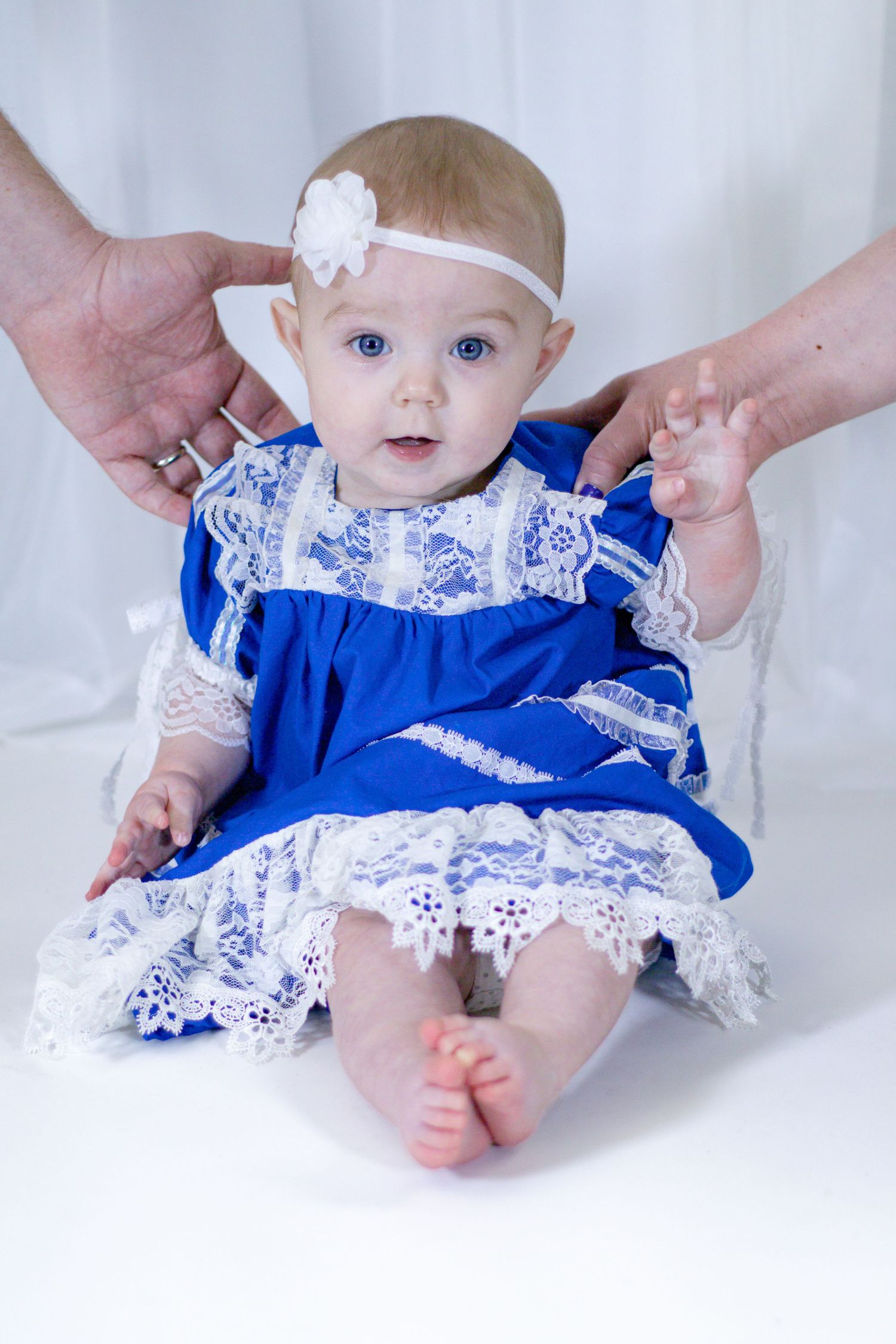 Blue-eyed infant in blue and white lace dress being supported by adult hands during photo session.