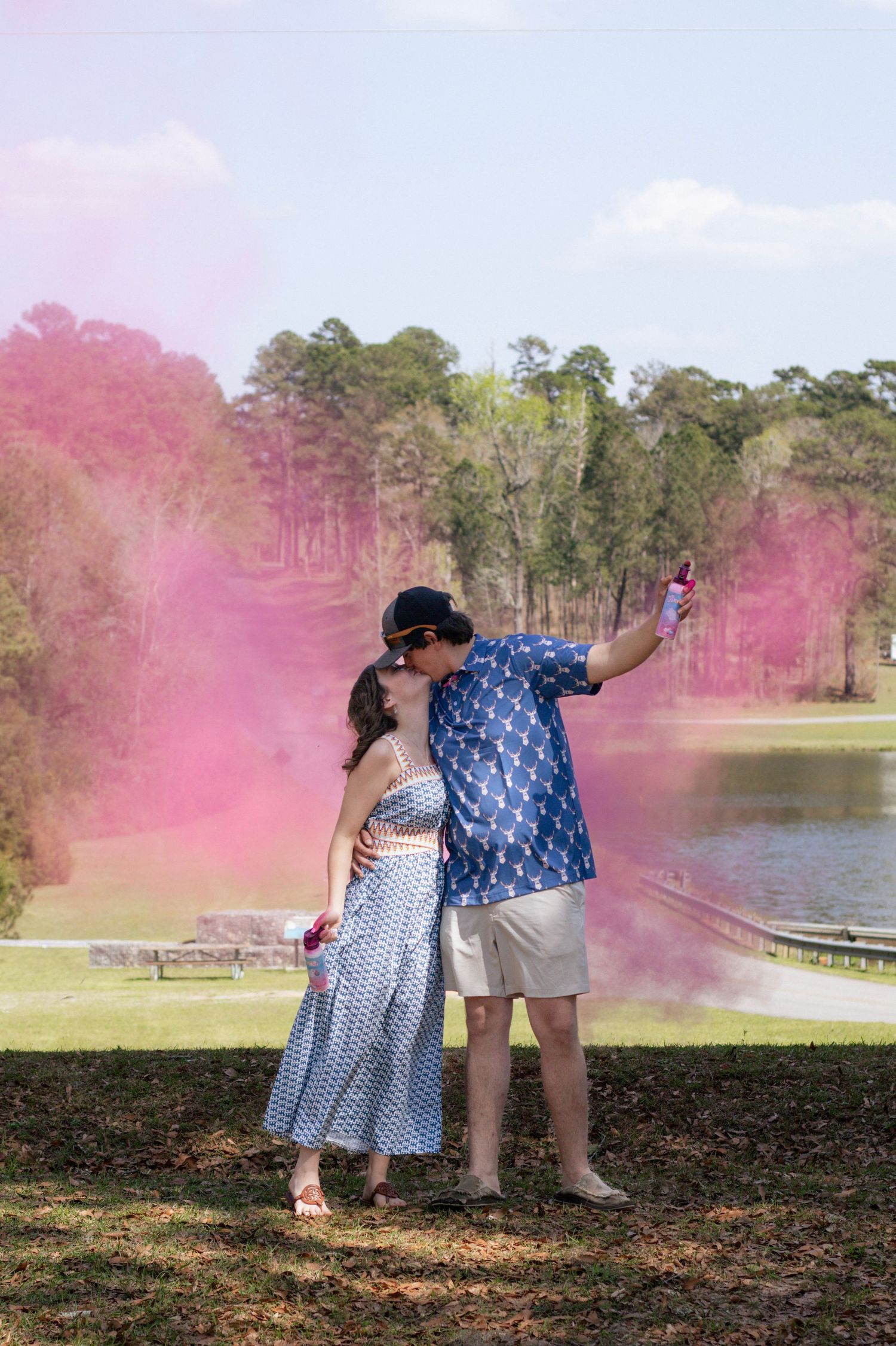 Couple kisses during pink smoke gender reveal outdoors by a lake.